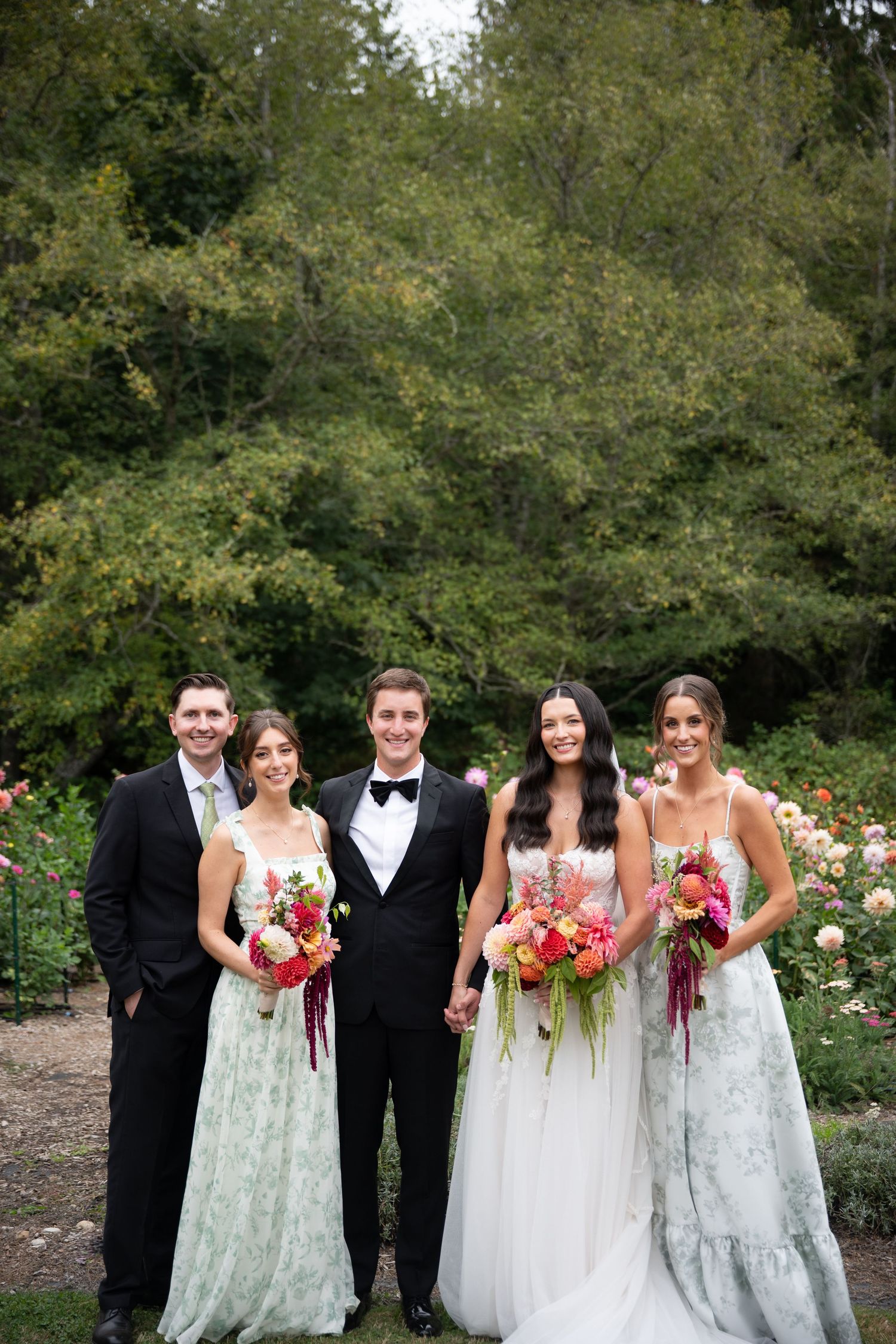 Wedding party posing outdoors with bride in white gown, bridesmaids in green floral dresses, and groomsmen in black tuxedos.