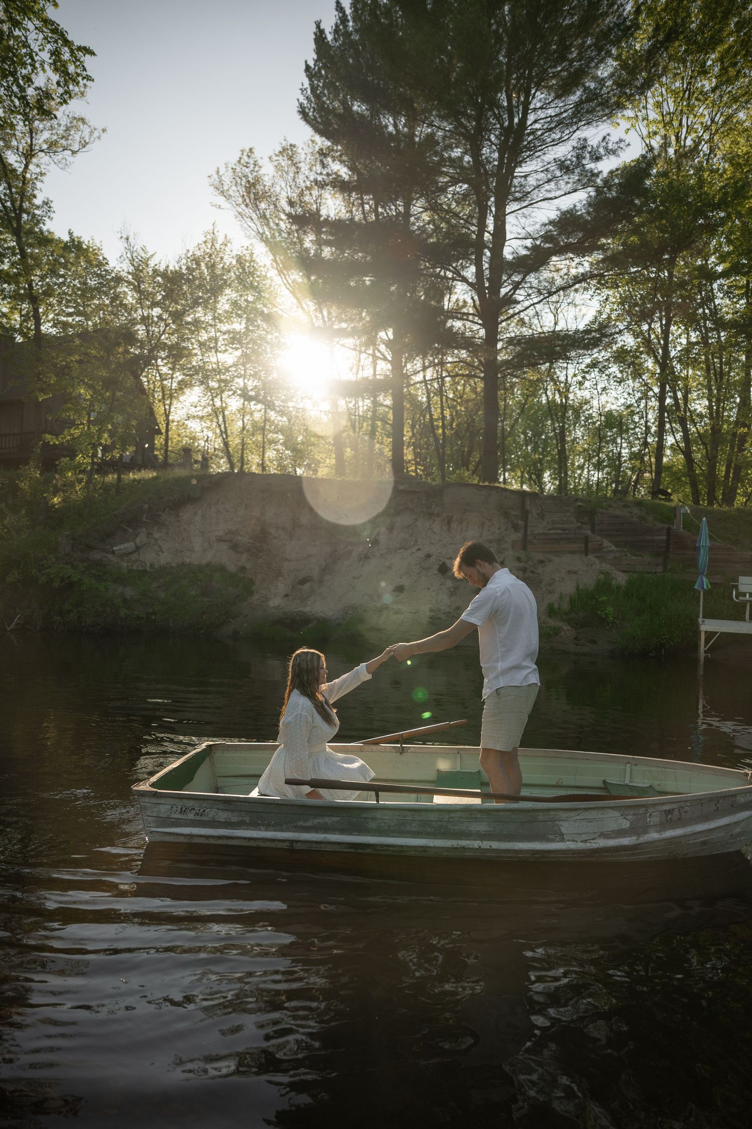 Couple holding hands on a rowboat at sunset, surrounded by lush trees reflecting on calm lake waters.