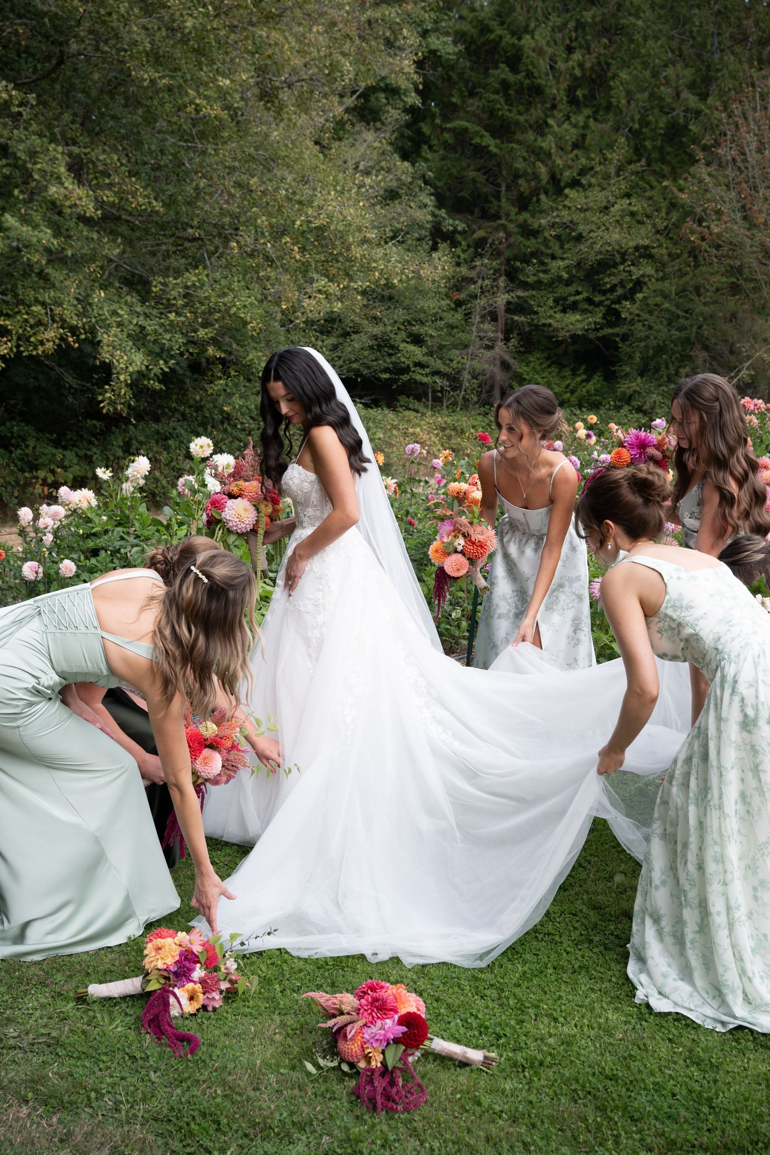 Bridesmaids in sage green dresses arrange the bride's white wedding gown train outdoors among colorful garden flowers.