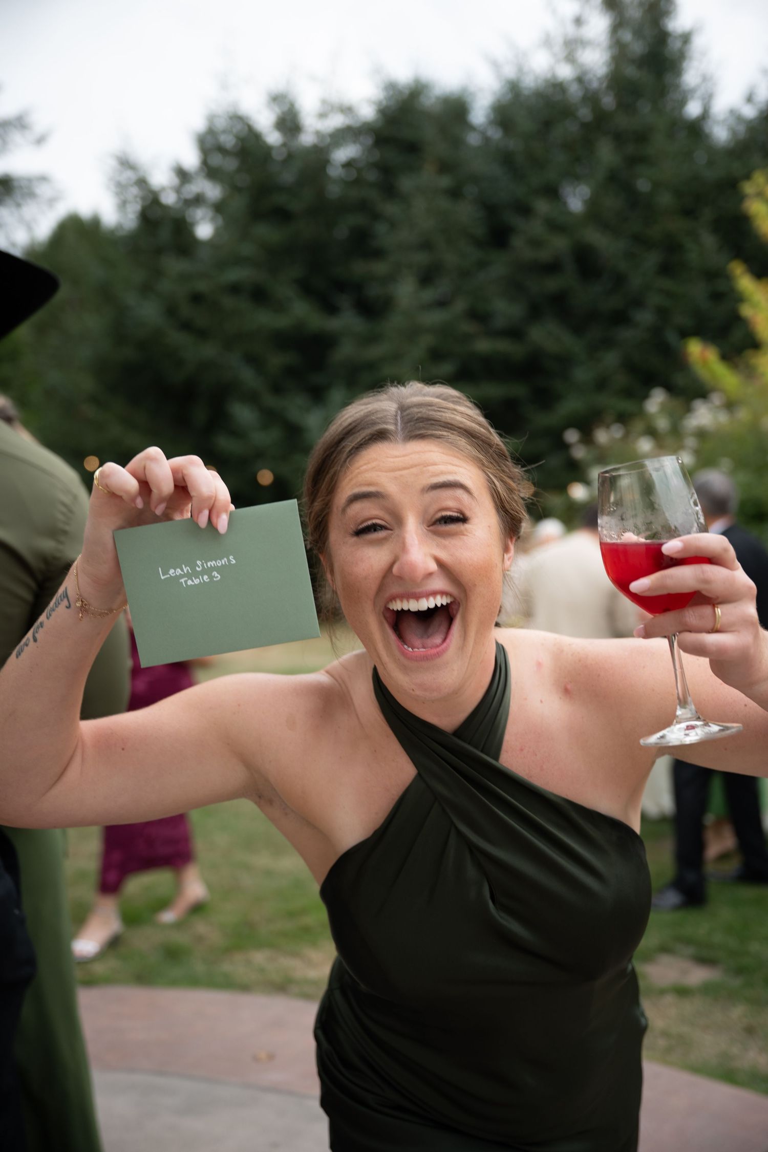 Excited woman in green halter dress holding seating card and red wine glass at outdoor wedding reception.