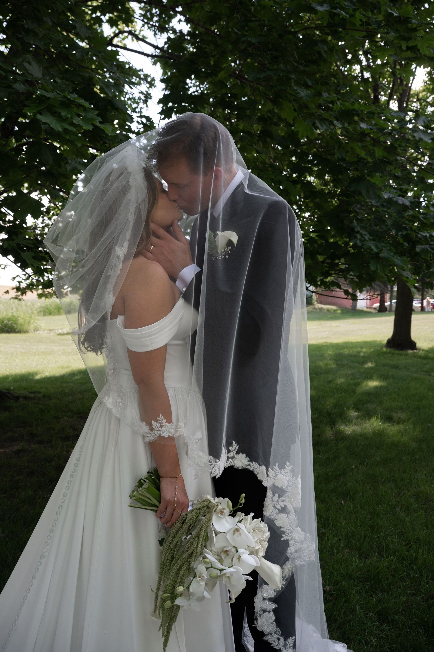 Bride and groom kissing under wedding veil, bride holding white bouquet in lush green garden.