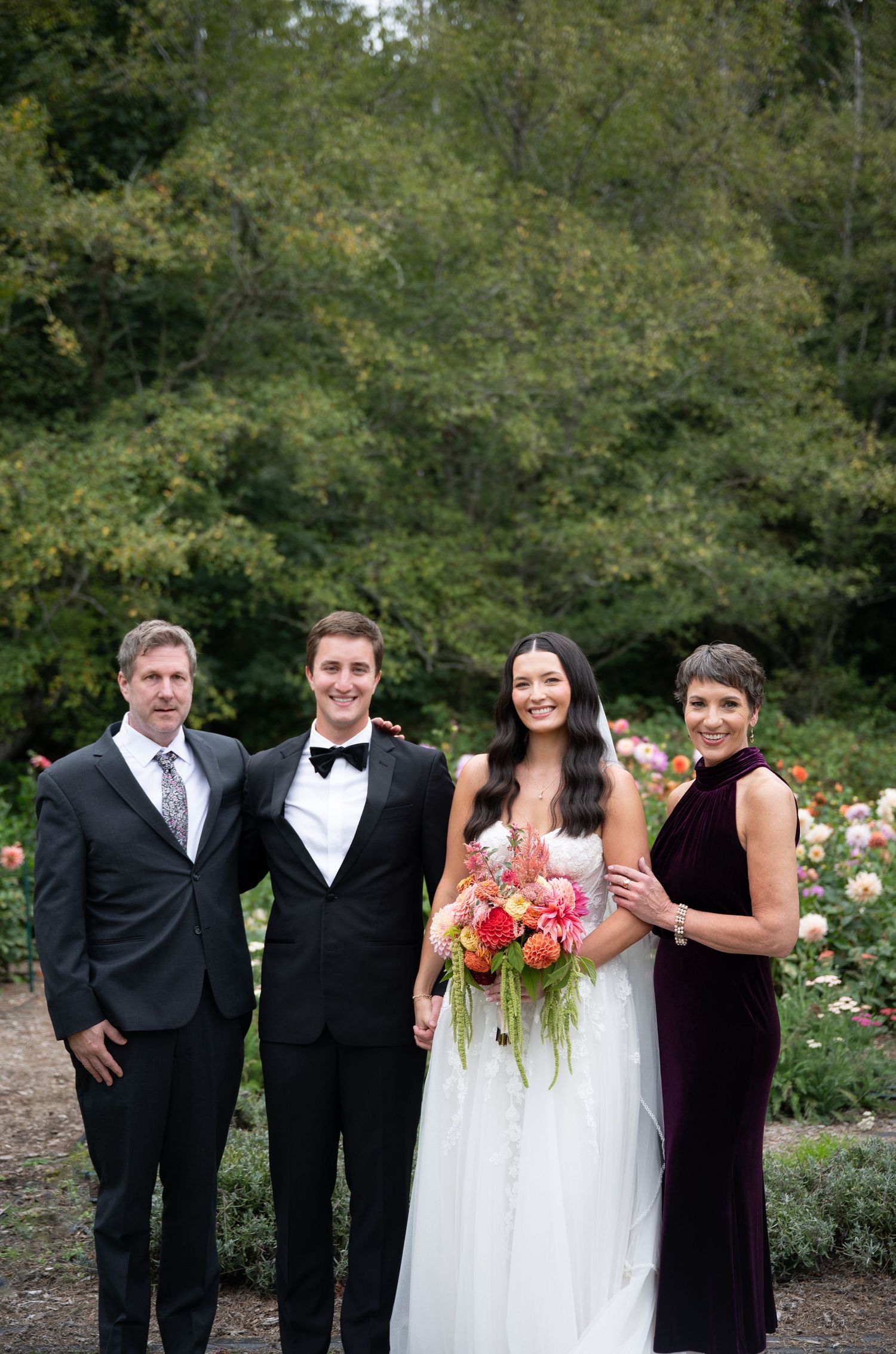 Wedding party family portrait with bride in white gown holding colorful bouquet flanked by parents in garden.
