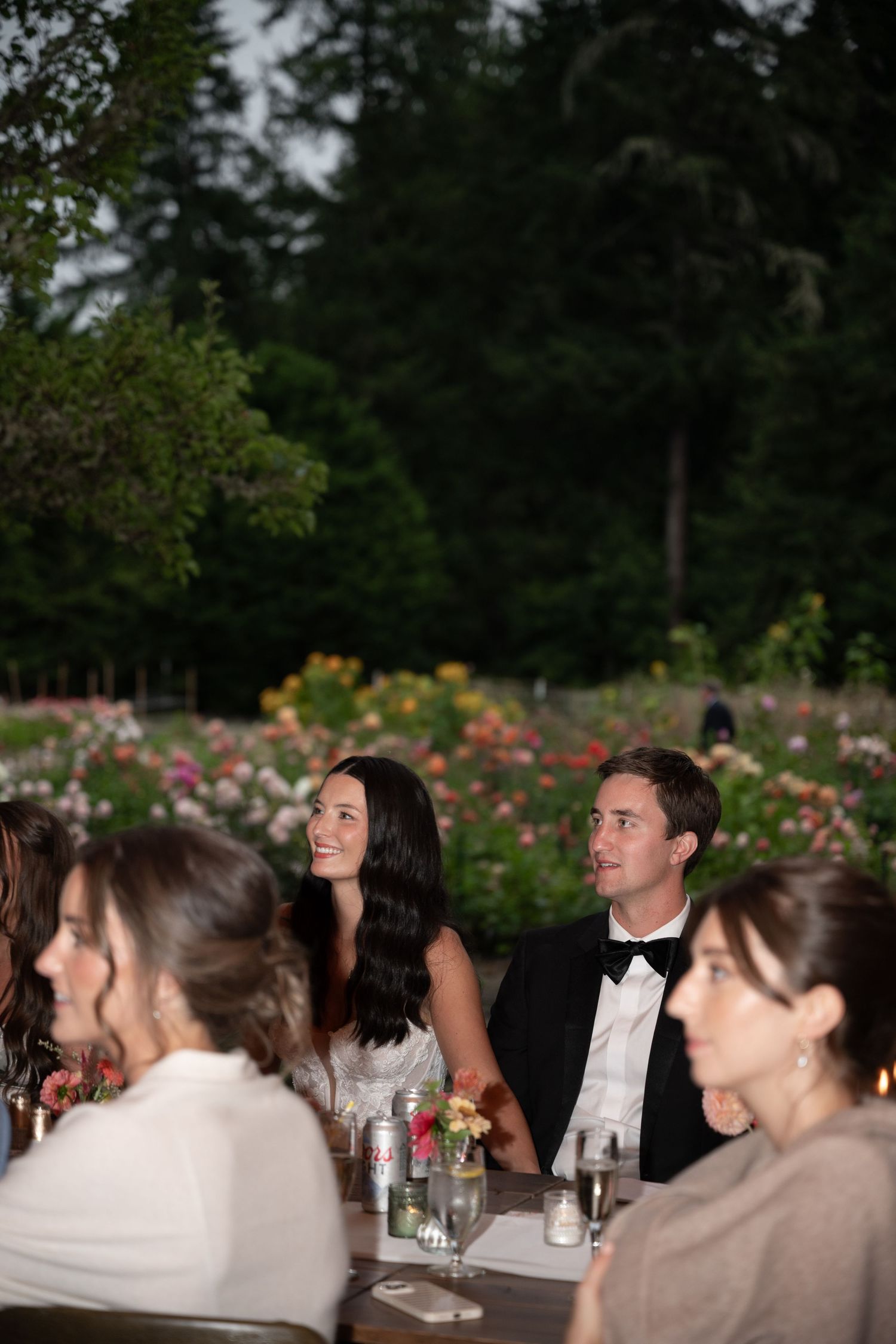 Bride and groom in formal attire sit at outdoor wedding reception table with rose garden backdrop at dusk.