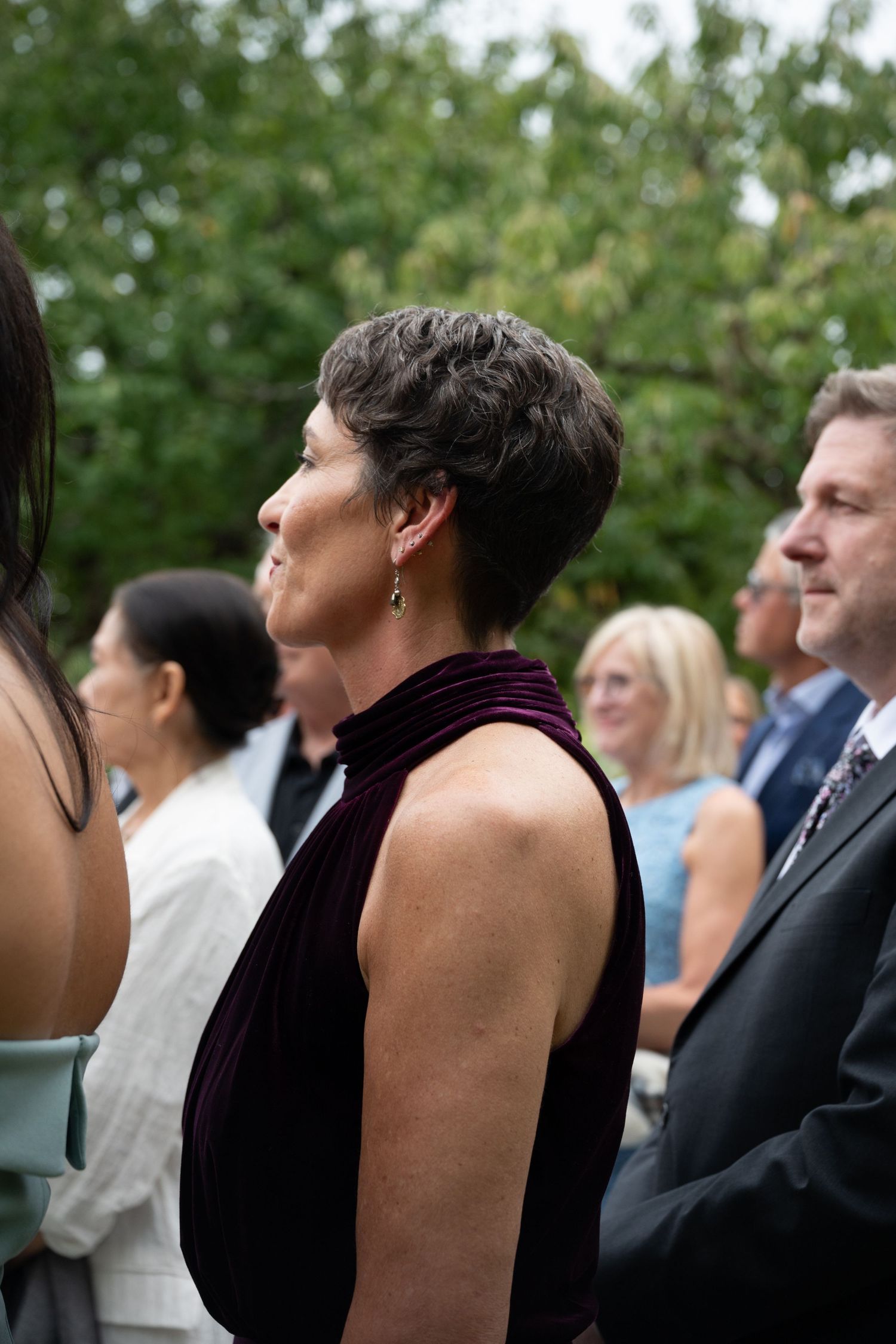 Woman in purple velvet dress standing among wedding ceremony guests outdoors.
