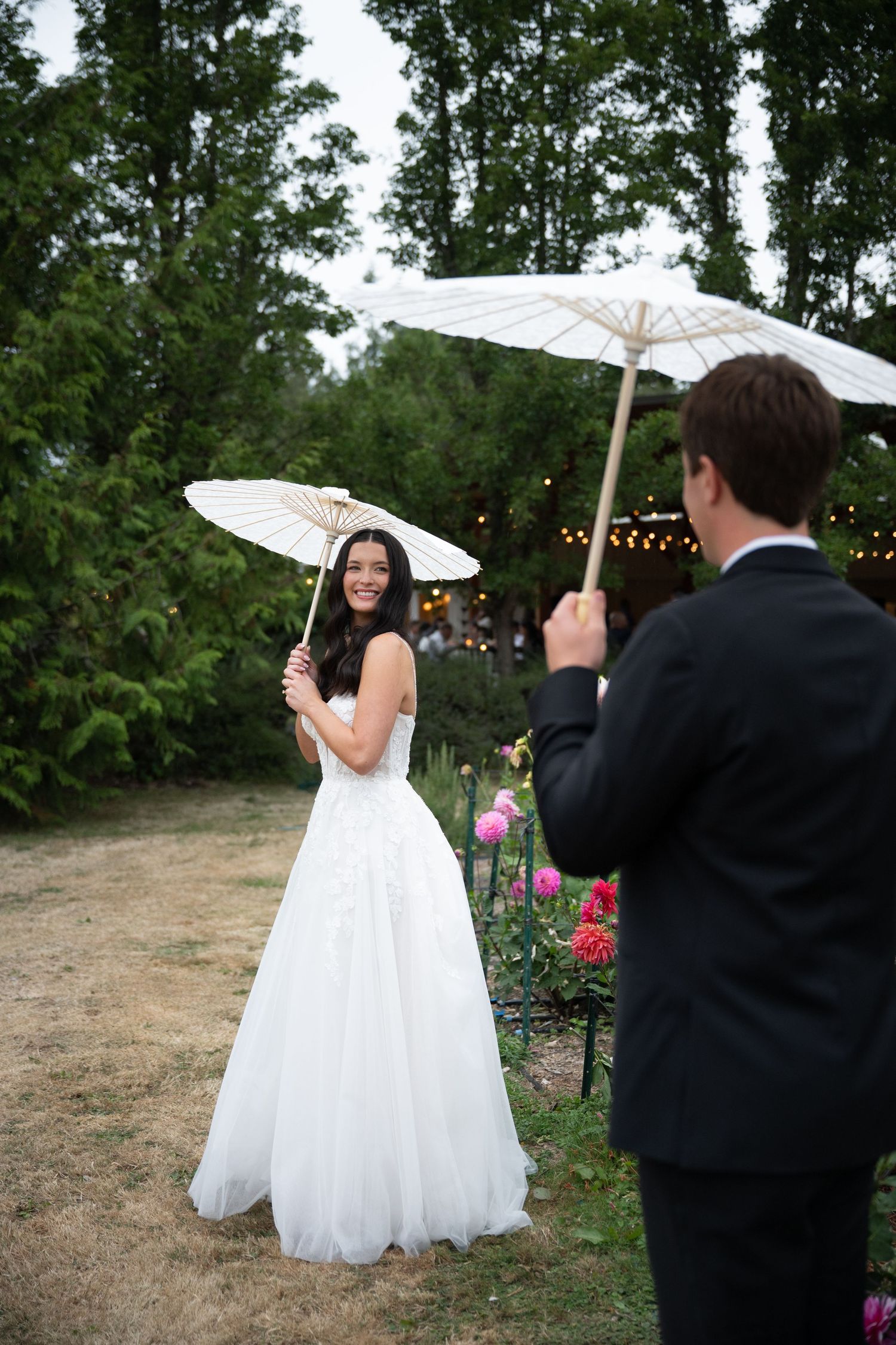Bride and groom hold white parasols while smiling at each other in garden setting.