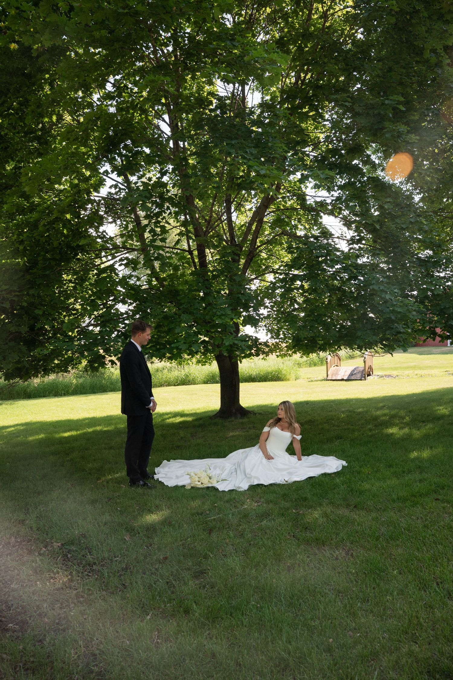 Bride in white gown sits on green grass while groom in black suit stands nearby under large shade trees.