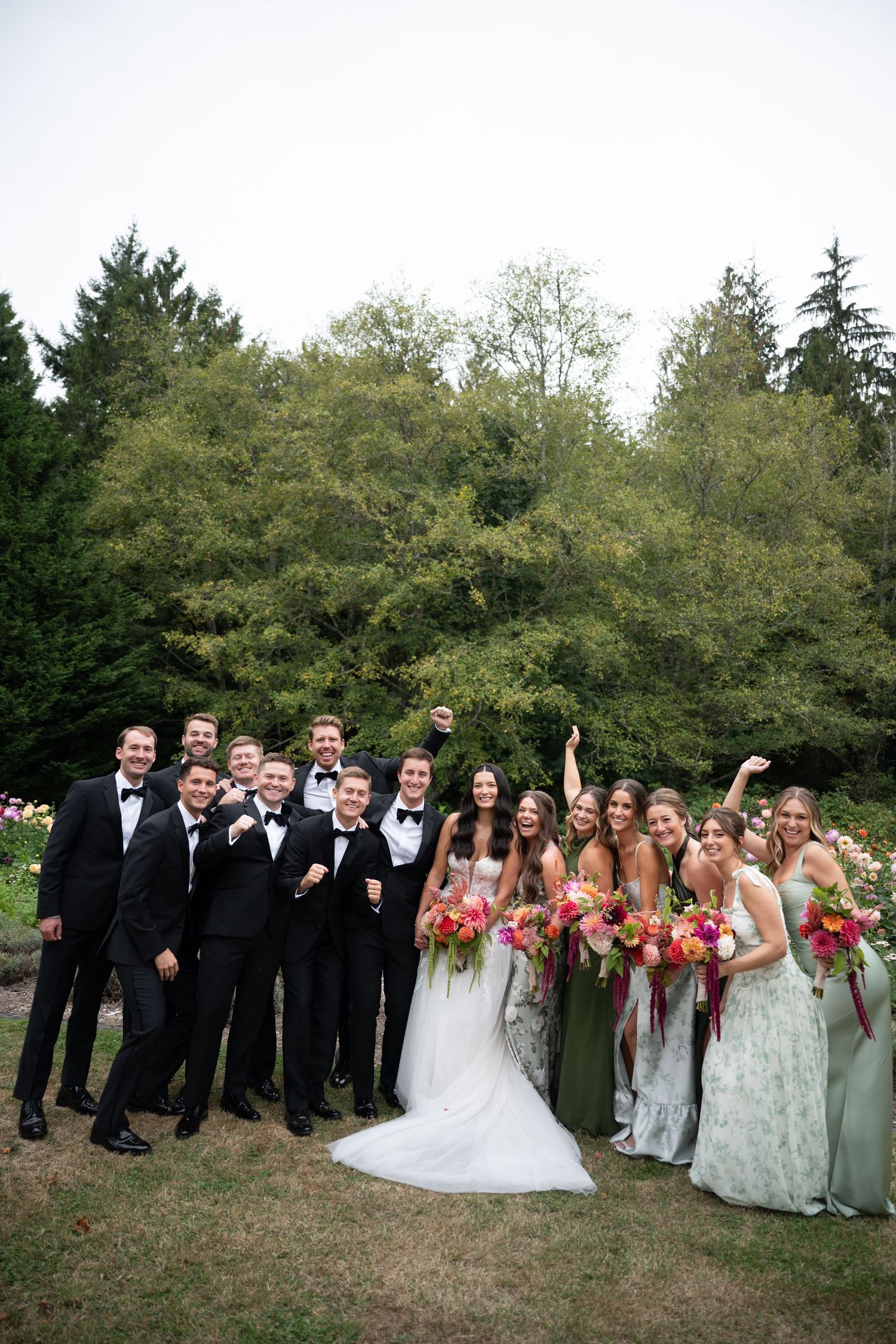 Wedding party poses outdoors, groomsmen in black tuxedos and bridesmaids in sage floral dresses holding vibrant bouquets.