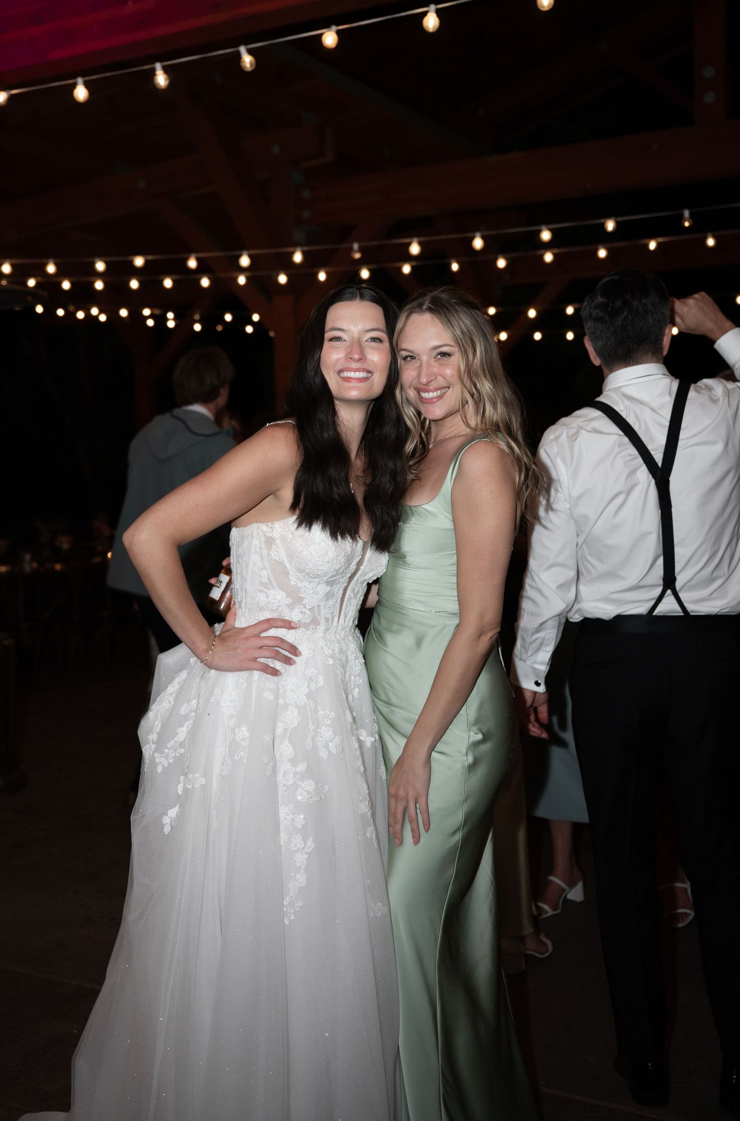Bride in white lace gown poses with bridesmaid in sage green dress at reception.