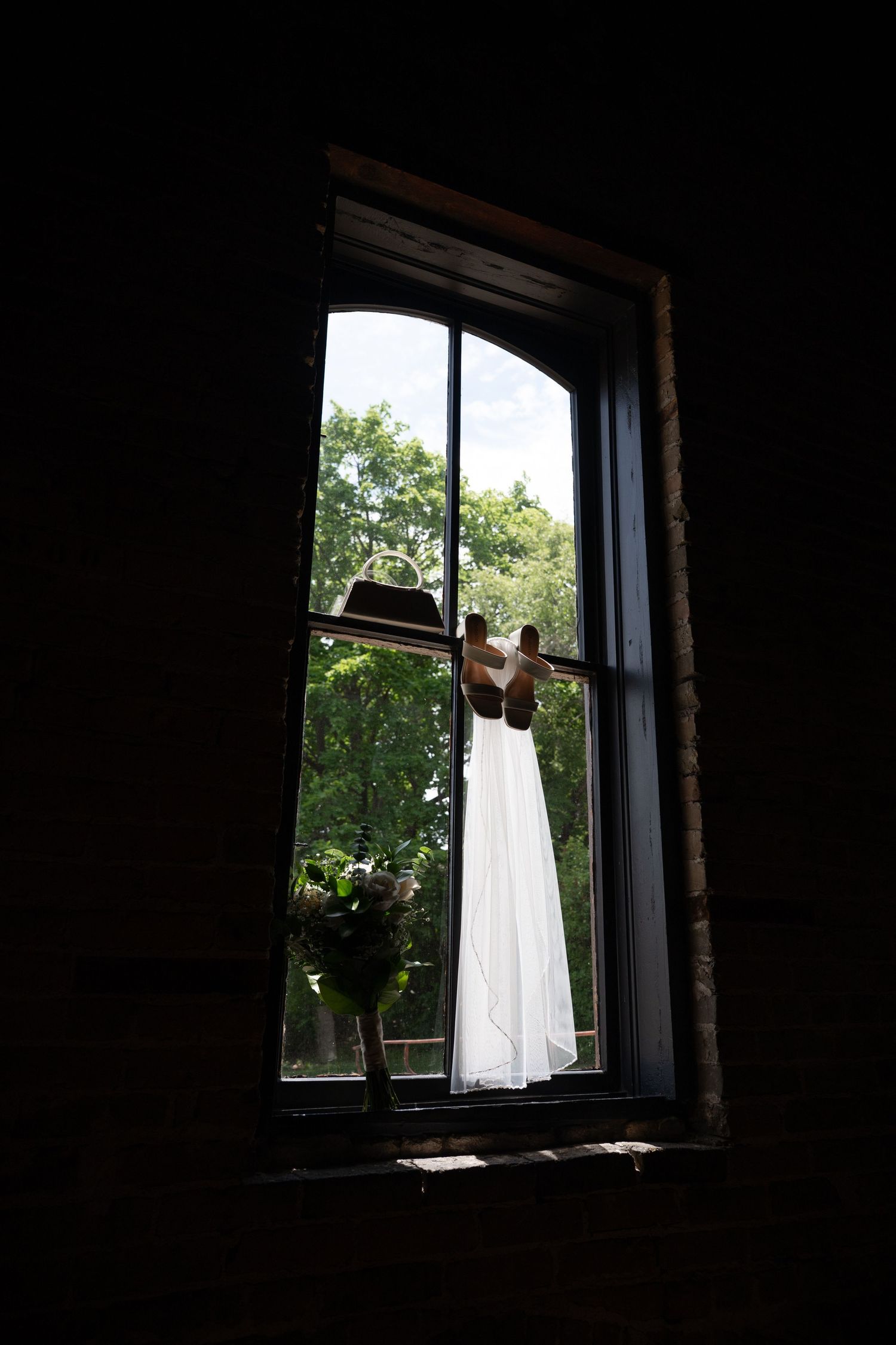 White wedding dress hanging in arched window with lush green trees and cowboy hat visible outside.