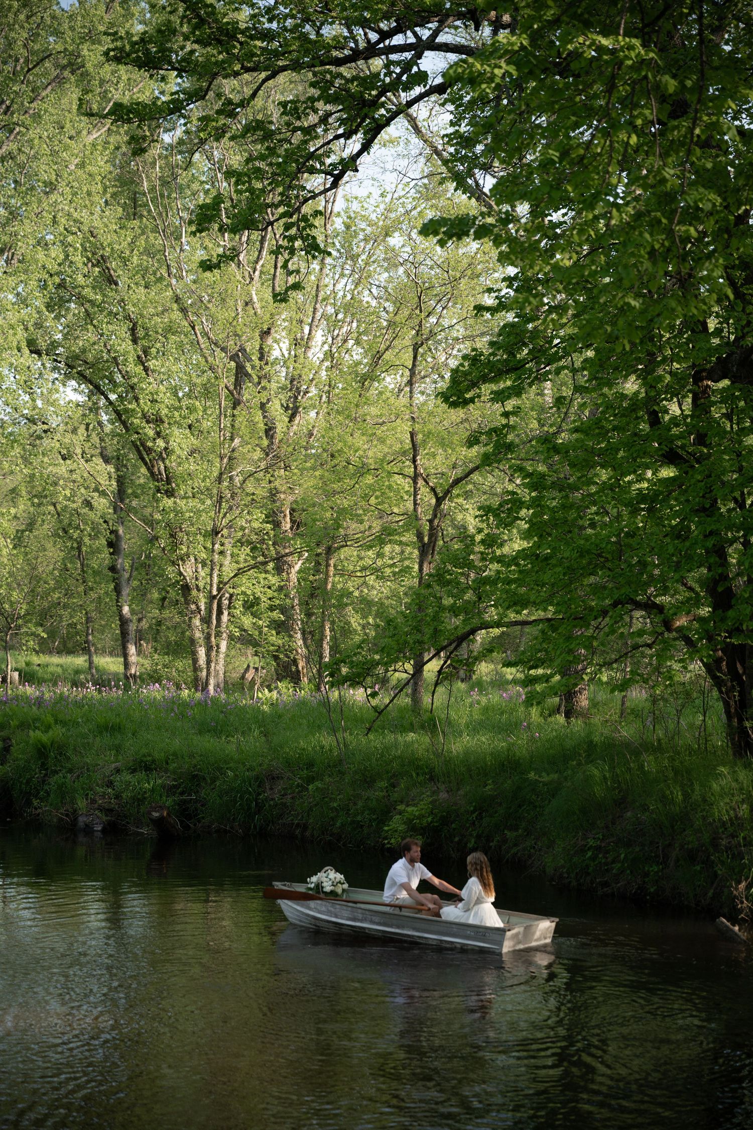 A couple in wedding attire sits in a small rowboat on a serene river surrounded by lush green trees.