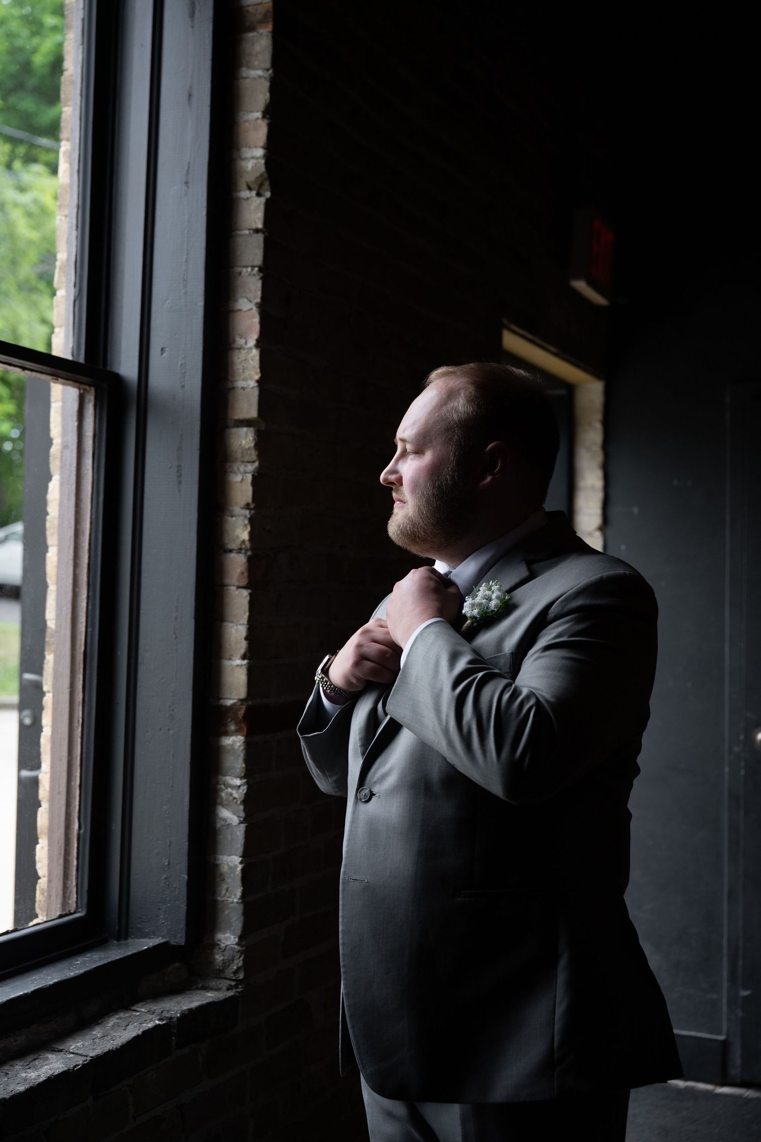 Handsome groom in gray suit adjusting tie while gazing out window in rustic brick venue.