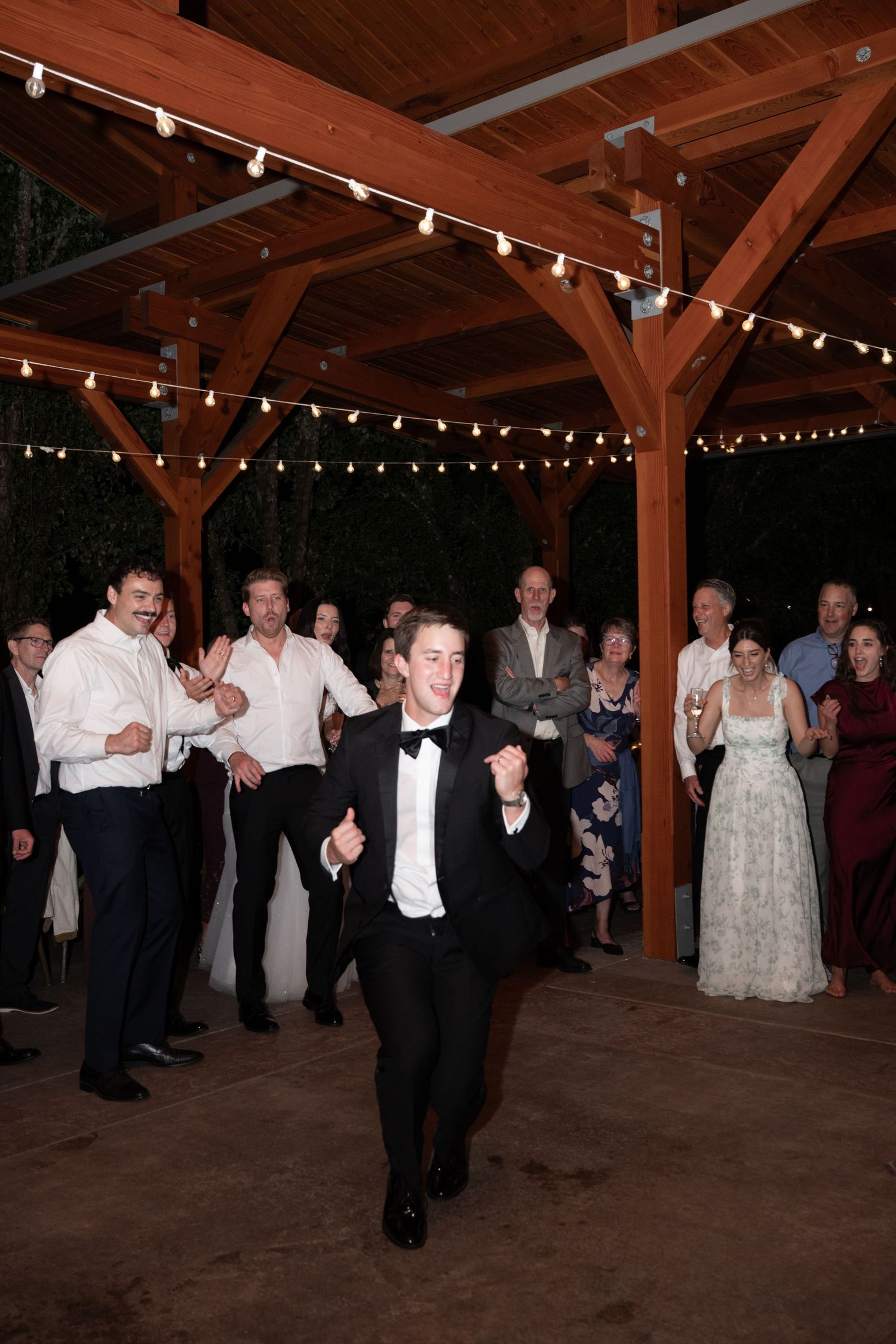 Groom dancing energetically under a wooden pergola with string lights as wedding guests cheer around him.