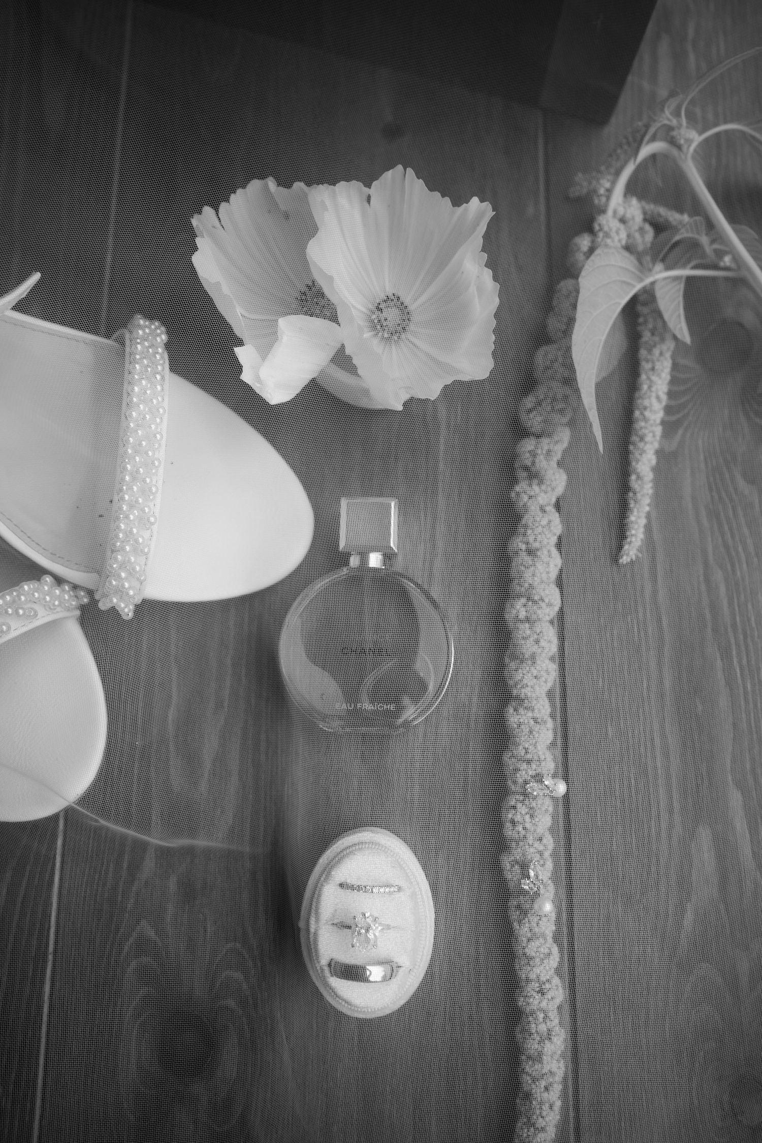 Black and white flat lay of bridal accessories including heels, perfume, rings, and flowers on wood.