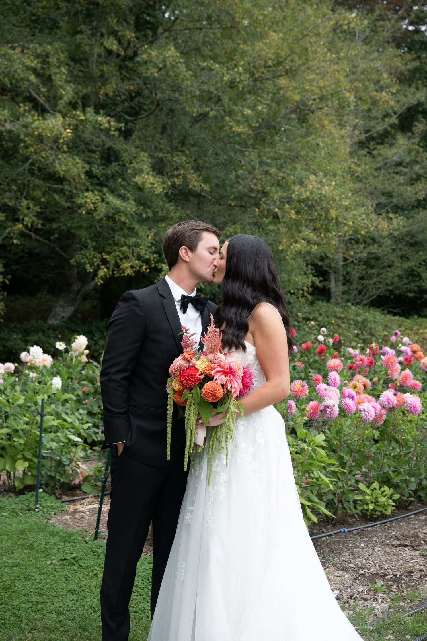 Bride and groom share a kiss in a lush garden surrounded by vibrant pink and coral flowers, bride holding a colorful bouquet.