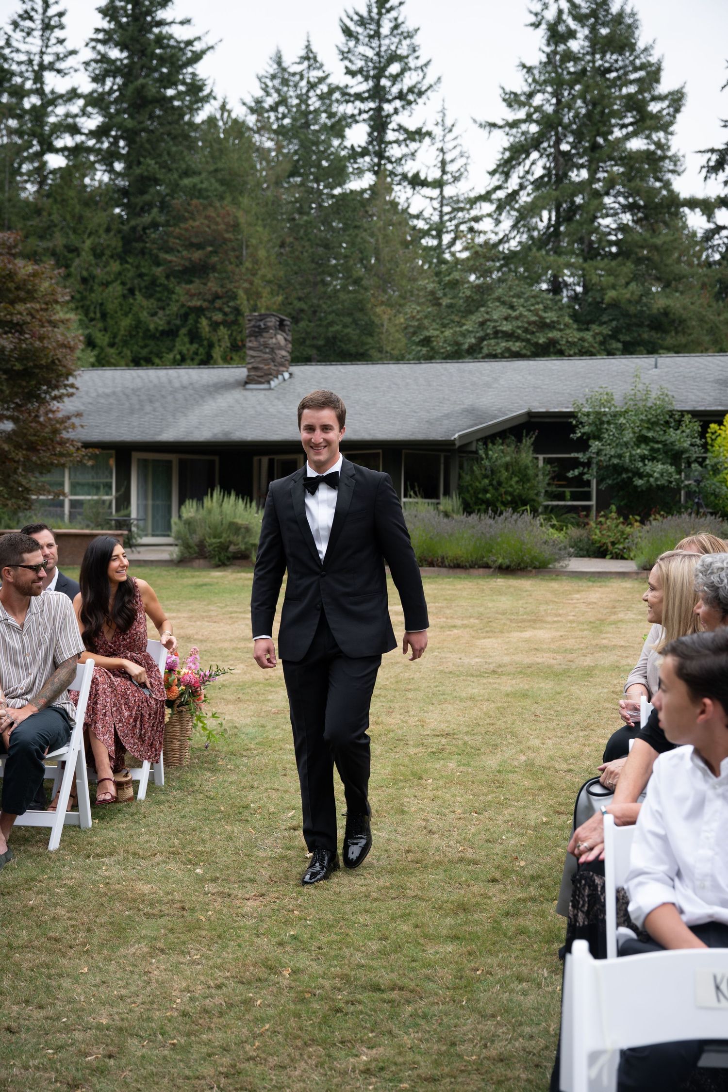 Groom in black tuxedo walks down outdoor wedding aisle flanked by seated guests on green lawn with trees behind.