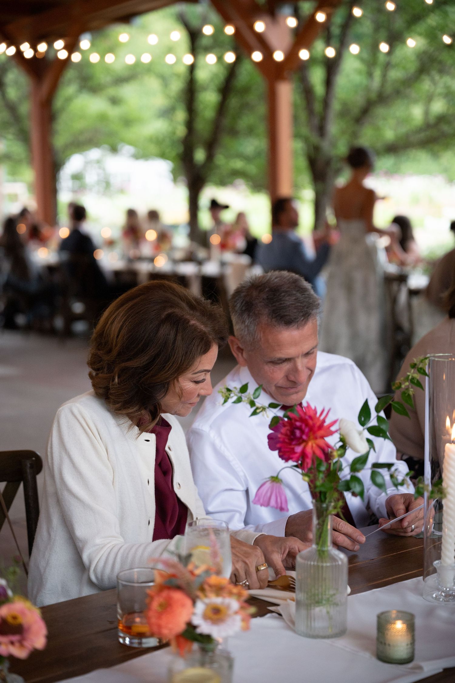 Couple shares a moment at wedding reception table with floral centerpieces under string lights.