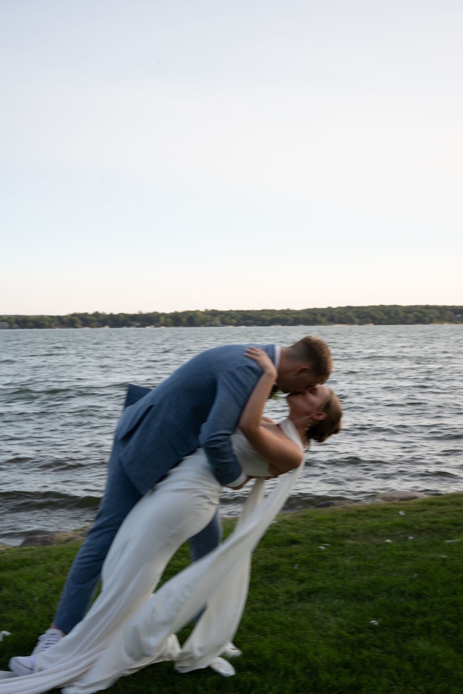 Couple sharing a romantic dip kiss by a lakeside at dusk, groom in blue suit and bride in flowing white dress.
