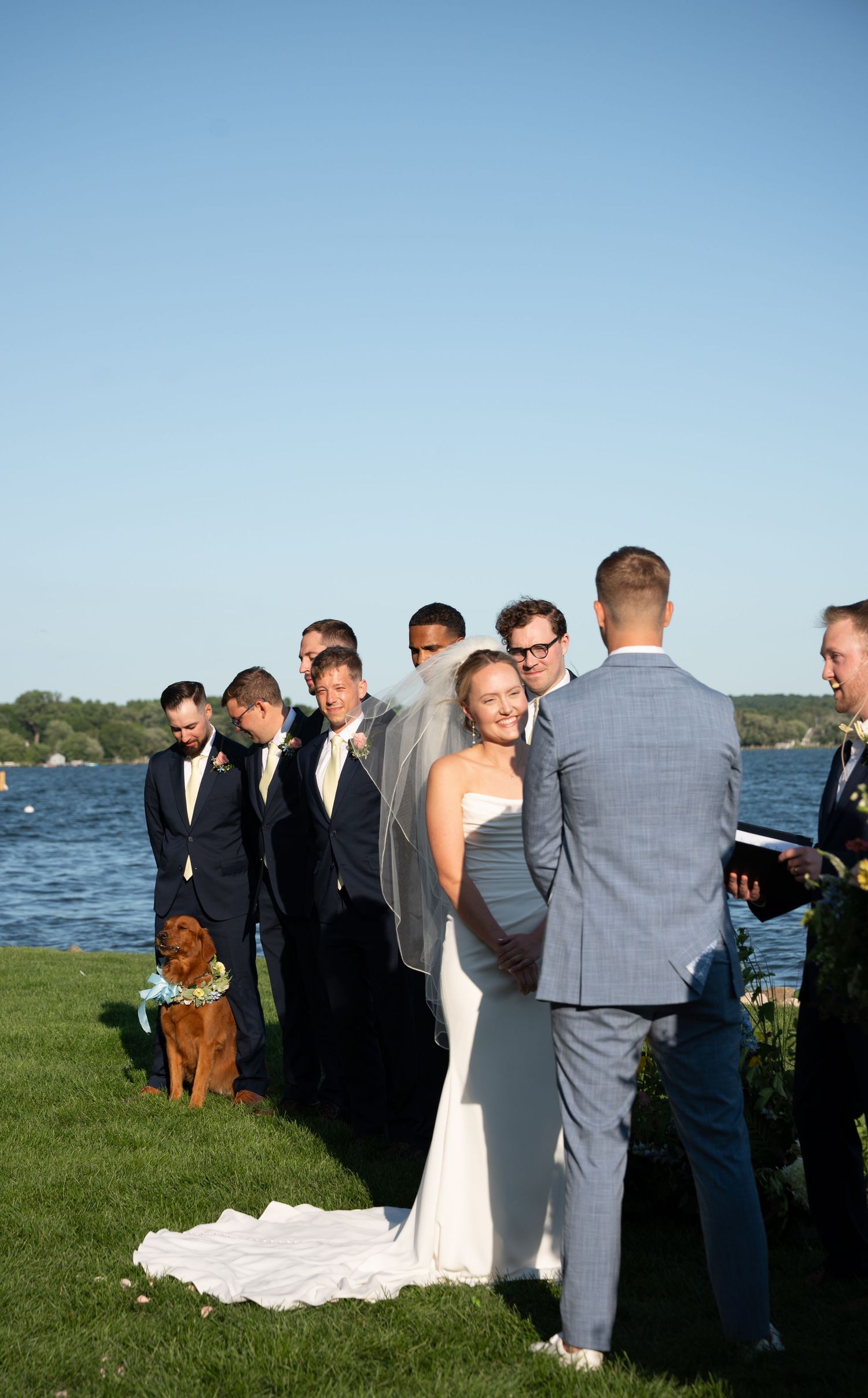 Outdoor waterfront wedding ceremony with bride in white gown, groom in gray suit, groomsmen in navy, and a dog in blue bow tie.