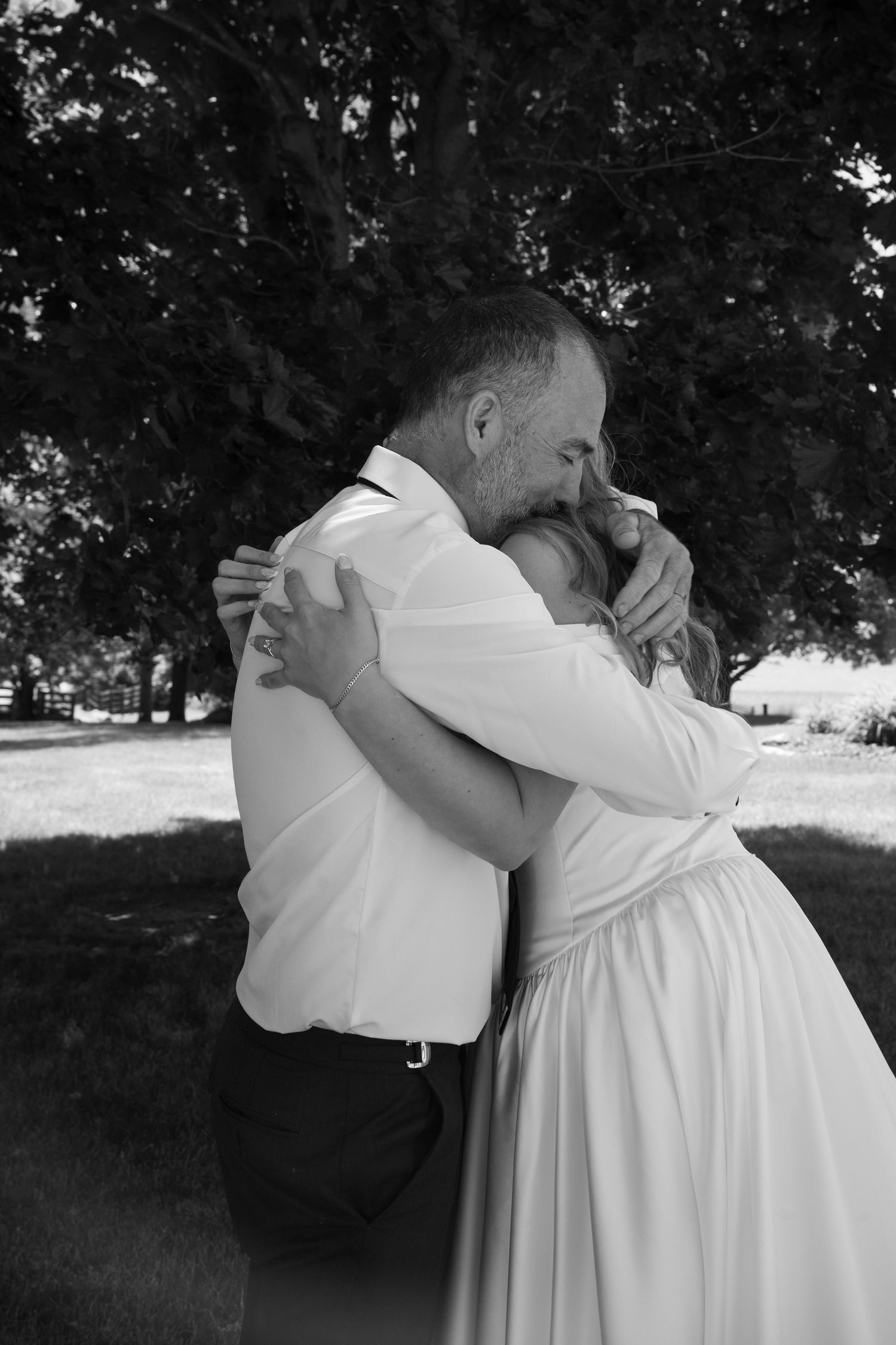 Black and white photo of a bride and groom sharing an emotional embrace outdoors under a large tree on their wedding day.