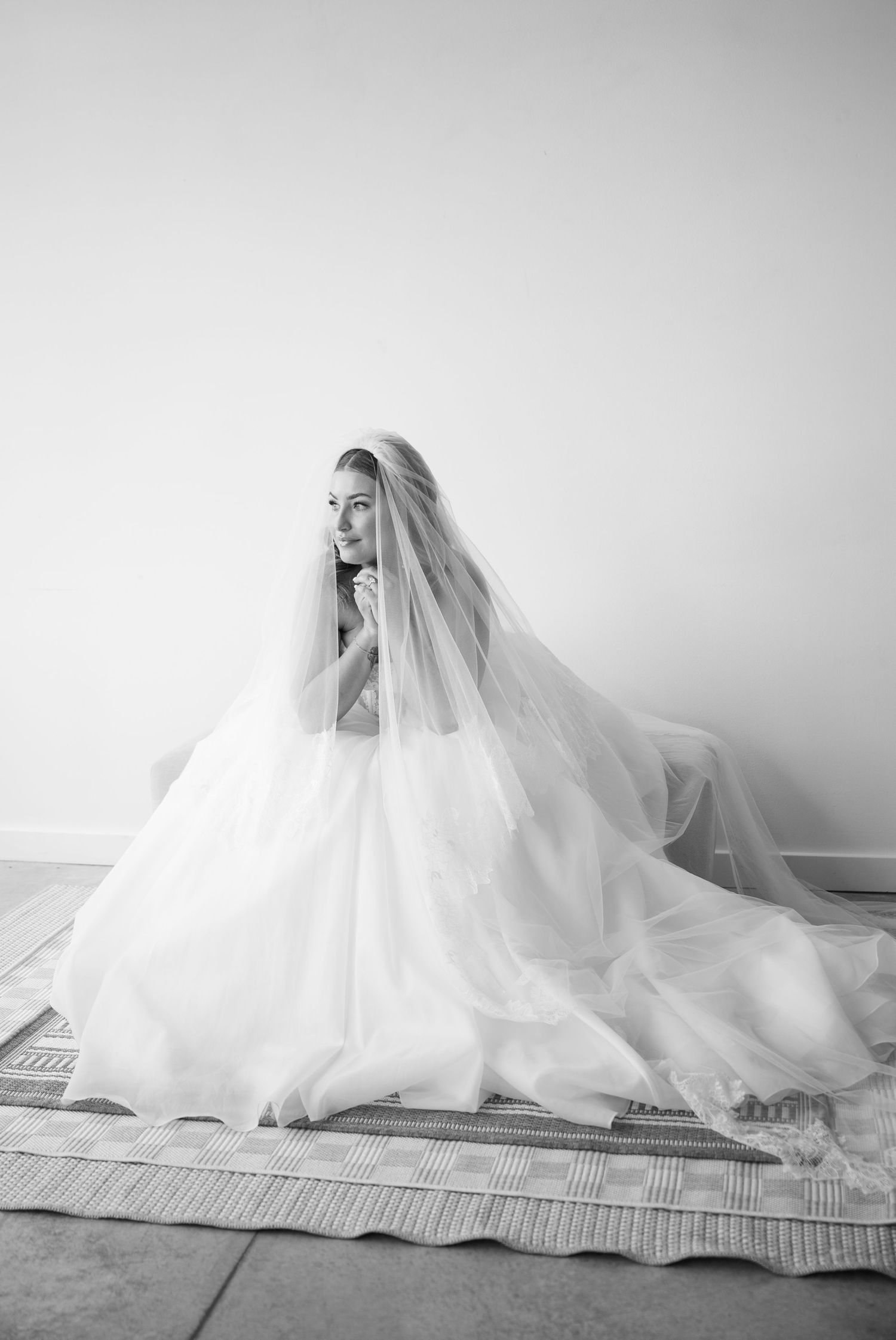 Black and white photo of a bride in a flowing wedding gown and veil seated gracefully on a rug.