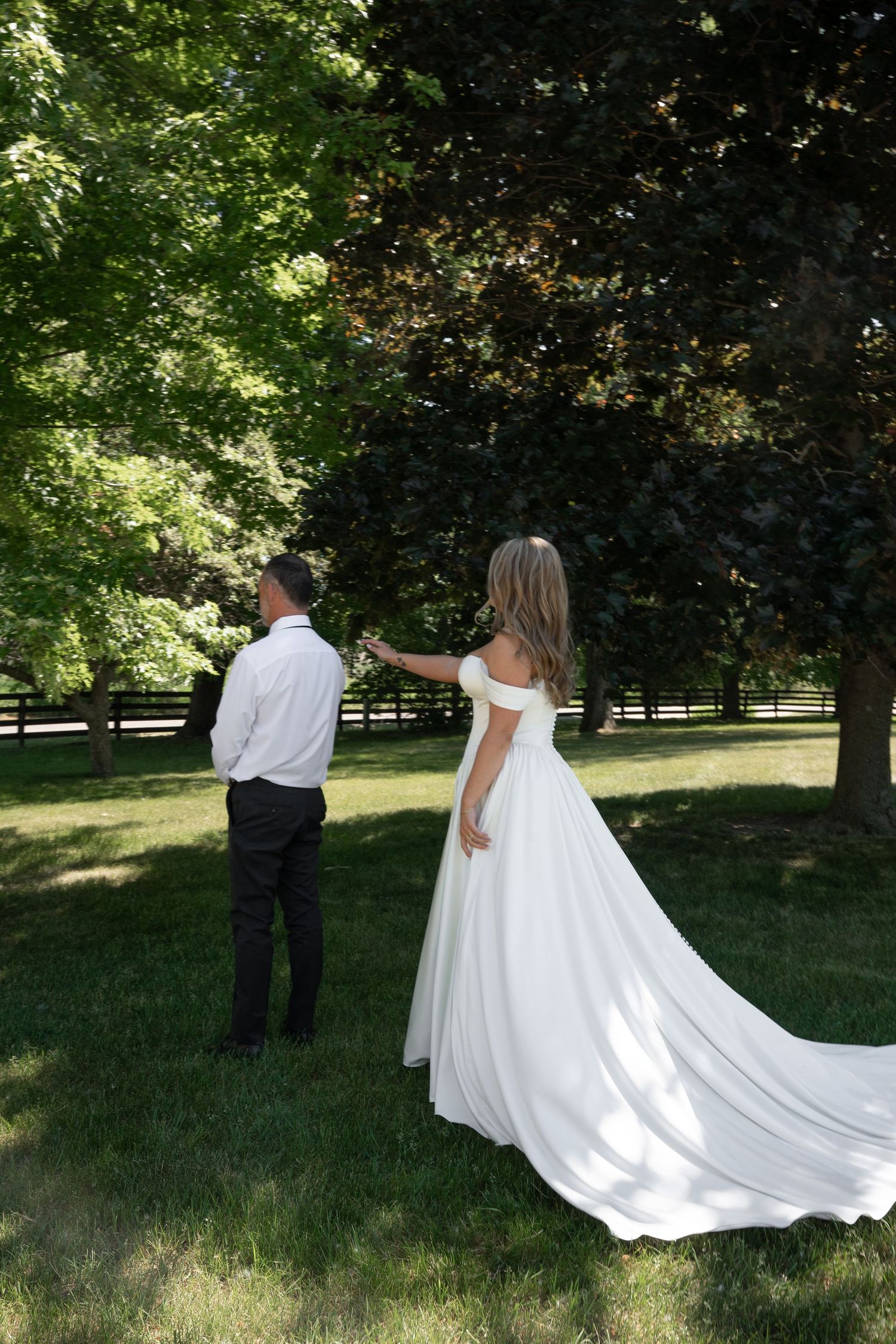 Bride in white off-shoulder gown with train taps groom on shoulder during outdoor first look among green trees.