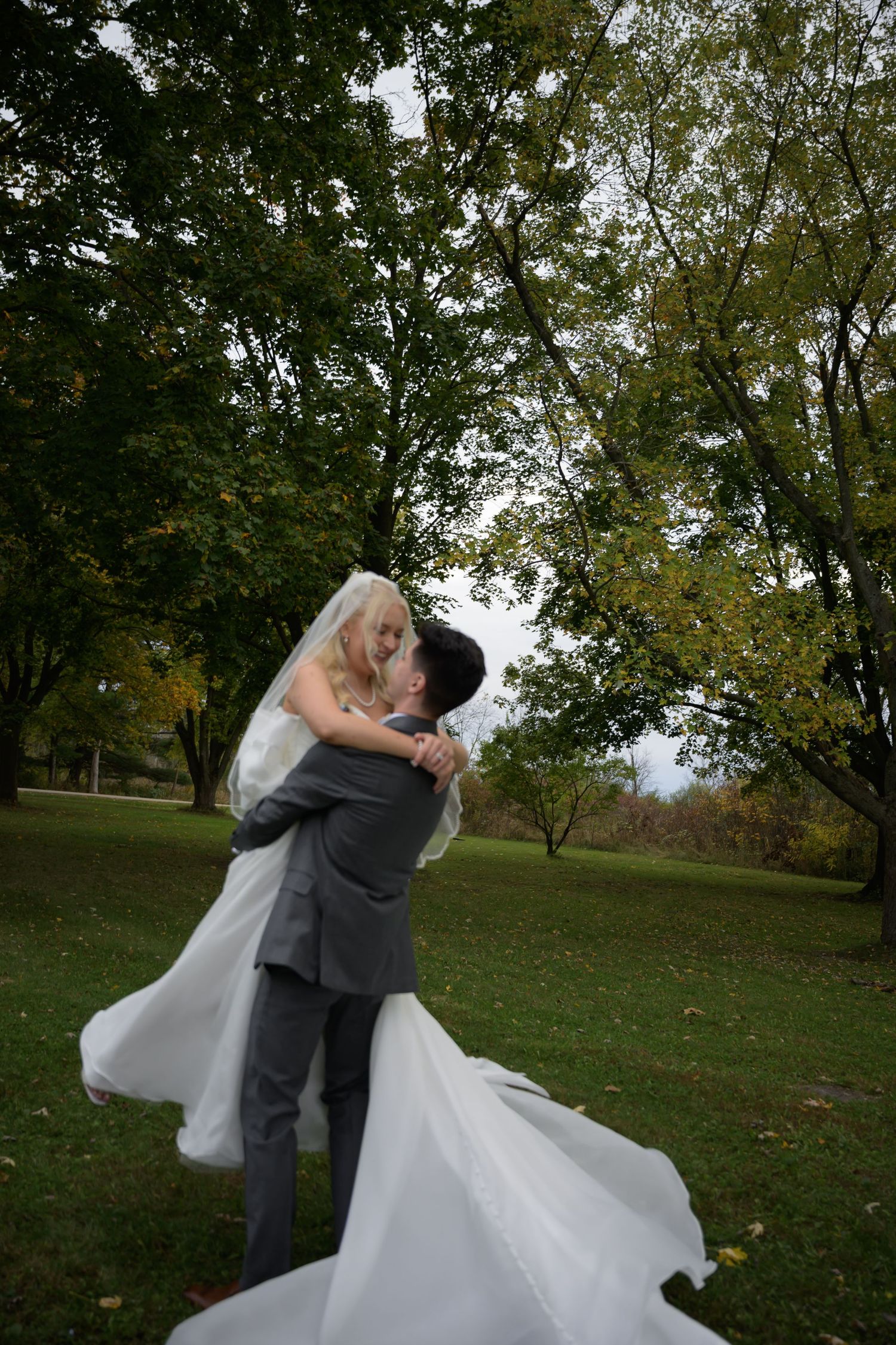 Groom lifting bride in white wedding dress and veil, sharing a romantic kiss in a lush green park setting.
