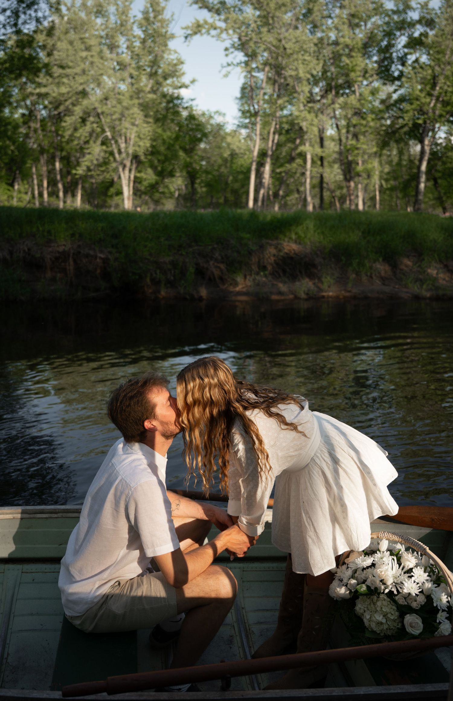 A romantic couple sharing a kiss on a wooden rowboat surrounded by lush green trees reflecting on a calm river.