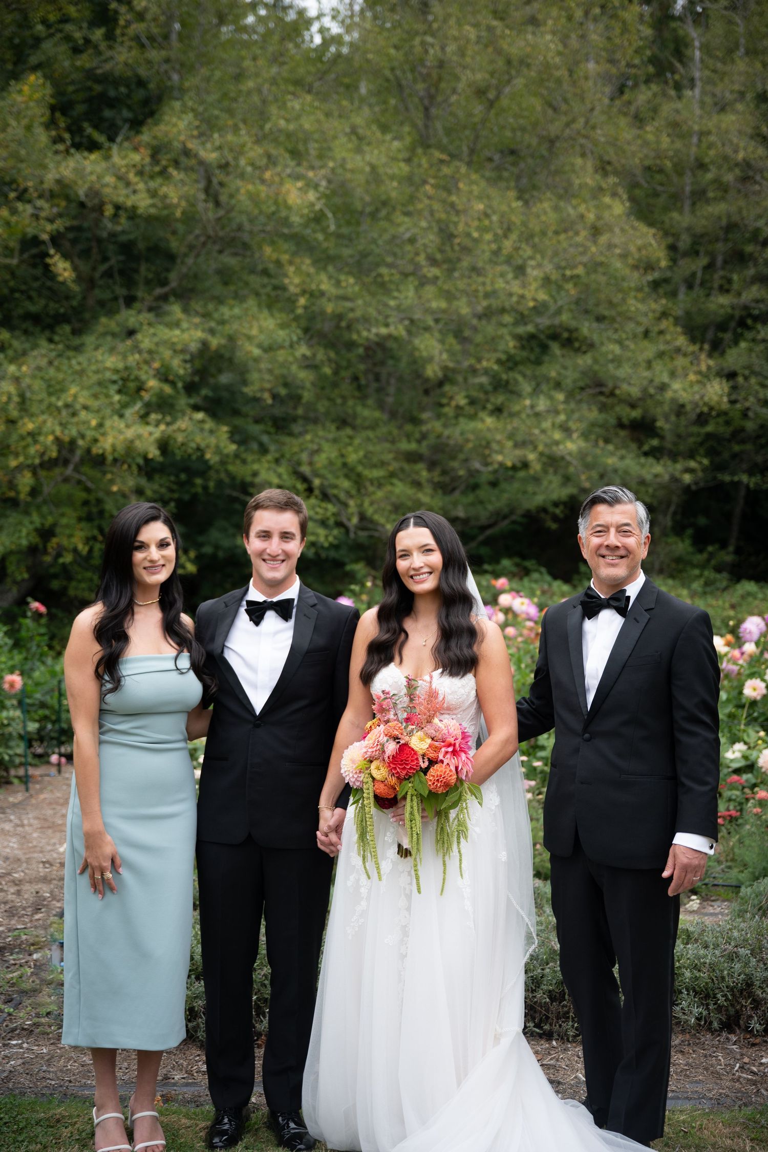 Bride in white gown and groom in tuxedo pose with family members in garden wedding portrait.