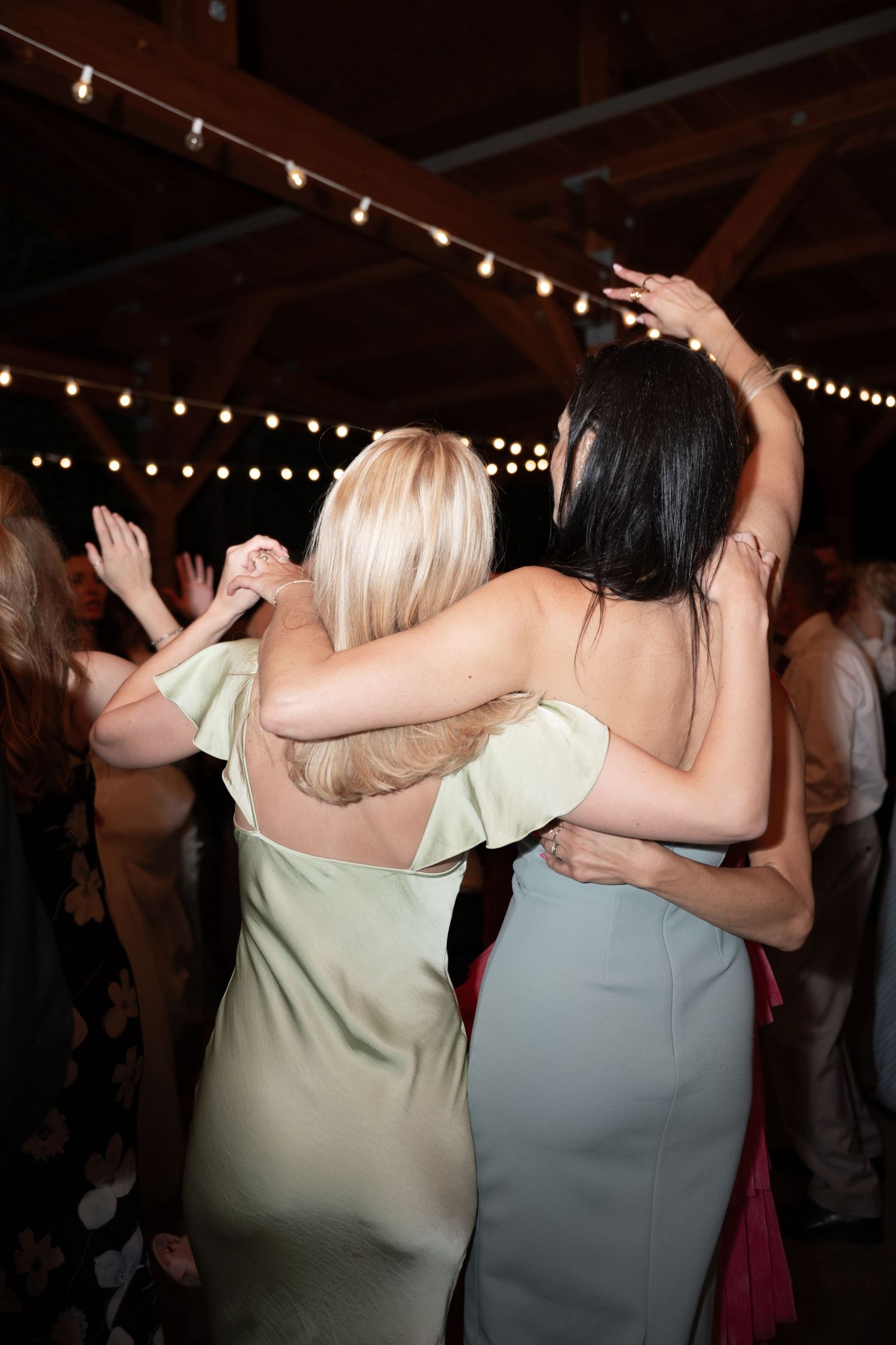 Two women in sage and black dresses dance together at wedding reception under string lights.