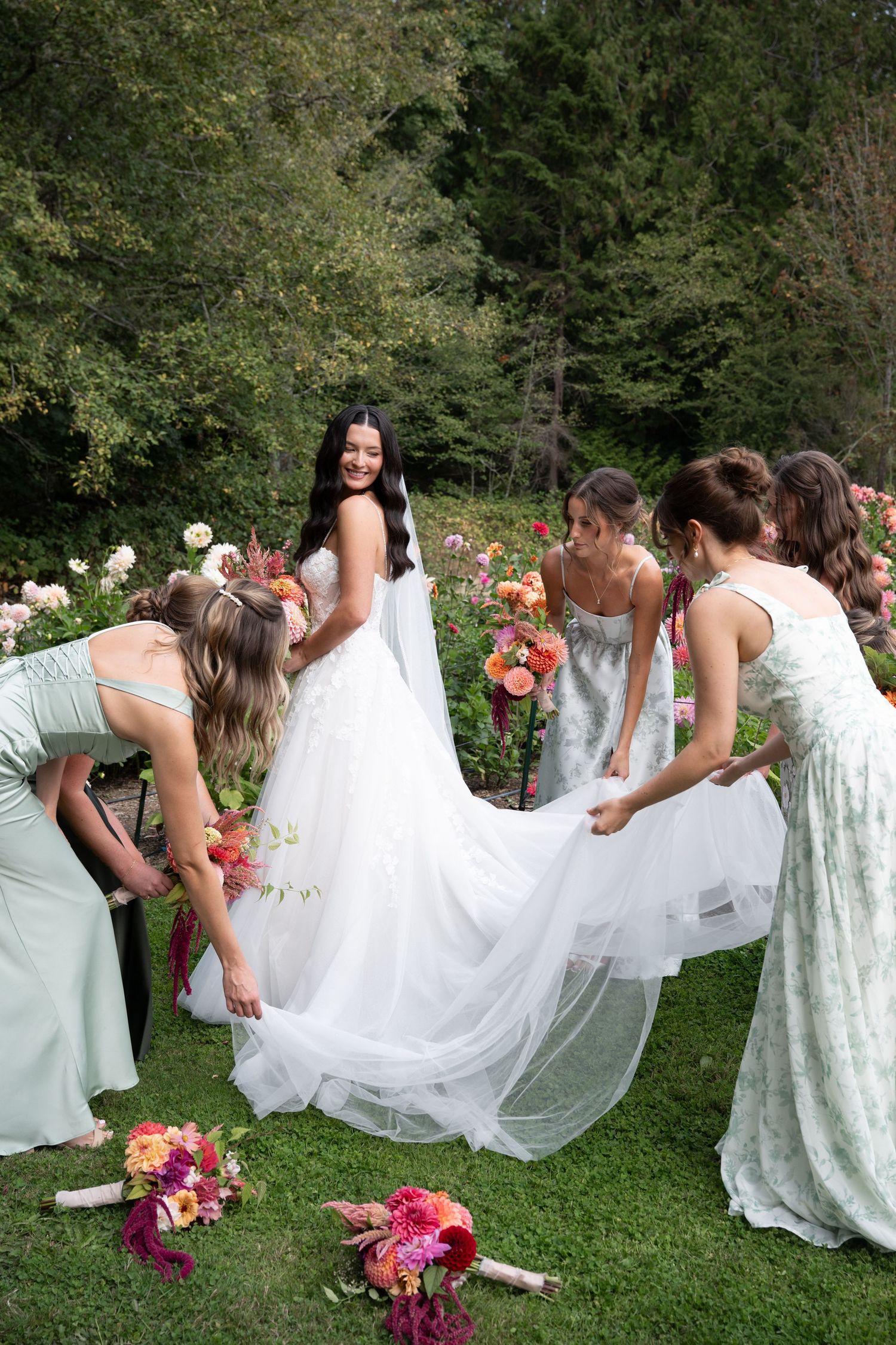 Bridesmaids in sage green dresses help adjust the bride's white gown in a lush garden with colorful floral bouquets.