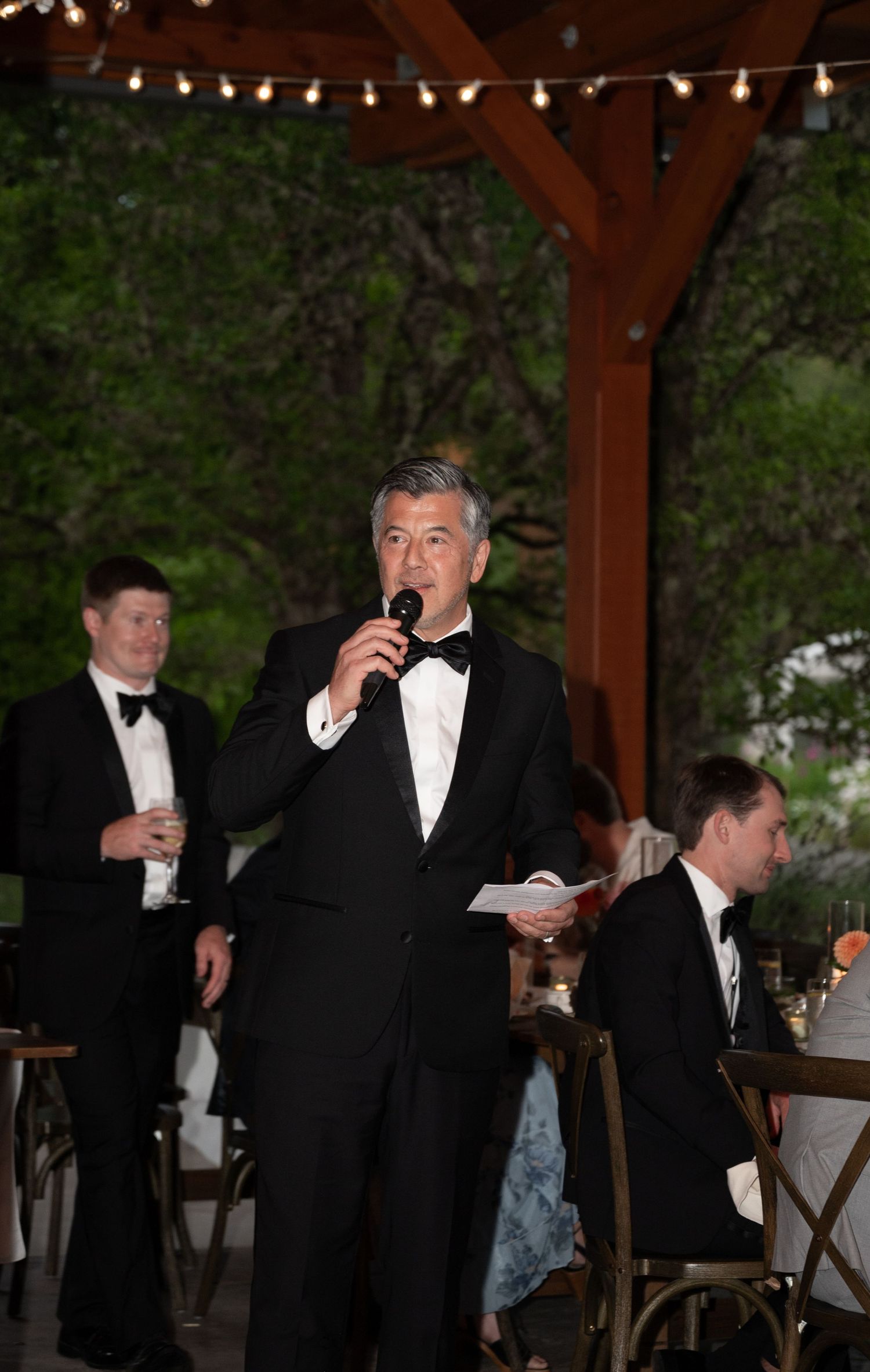 Man in tuxedo delivers wedding speech into microphone at elegant outdoor reception under string lights.