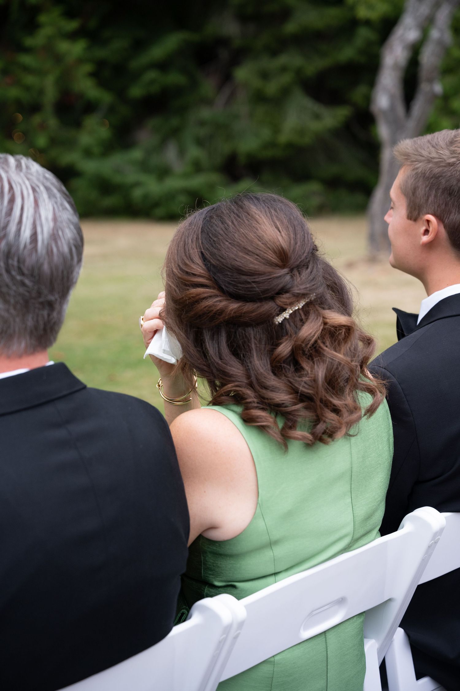 Emotional wedding guest in green dress wipes tears while seated at outdoor garden ceremony.