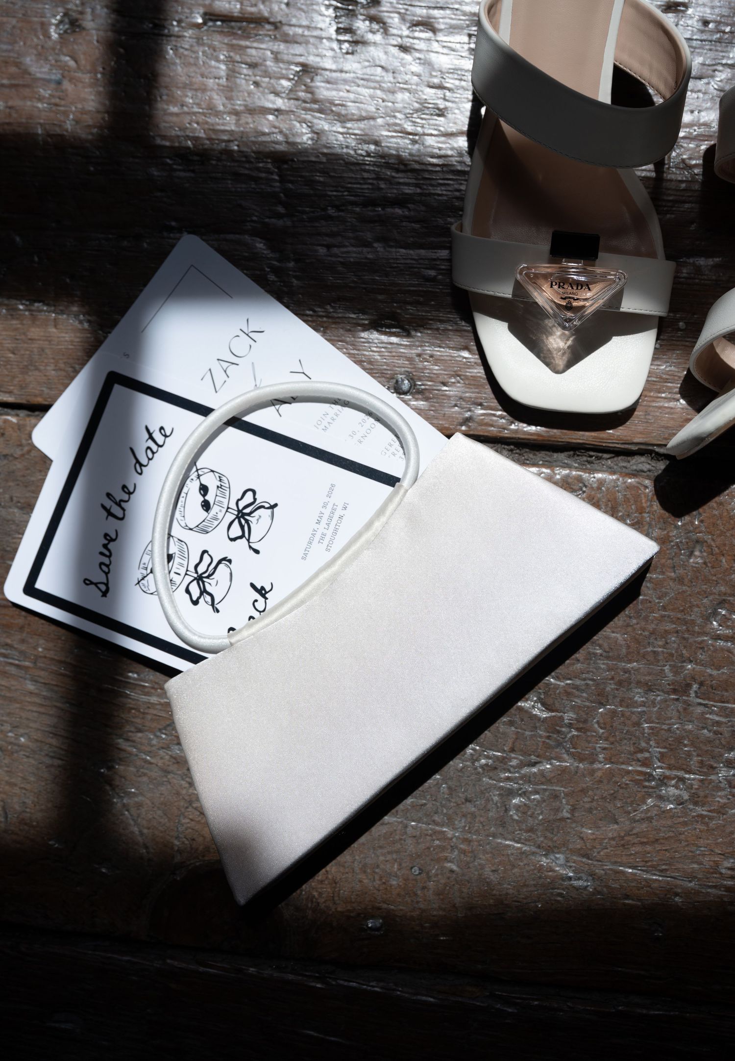Wedding save the date cards beside a white clutch purse and bridal heels on a rustic wooden surface.
