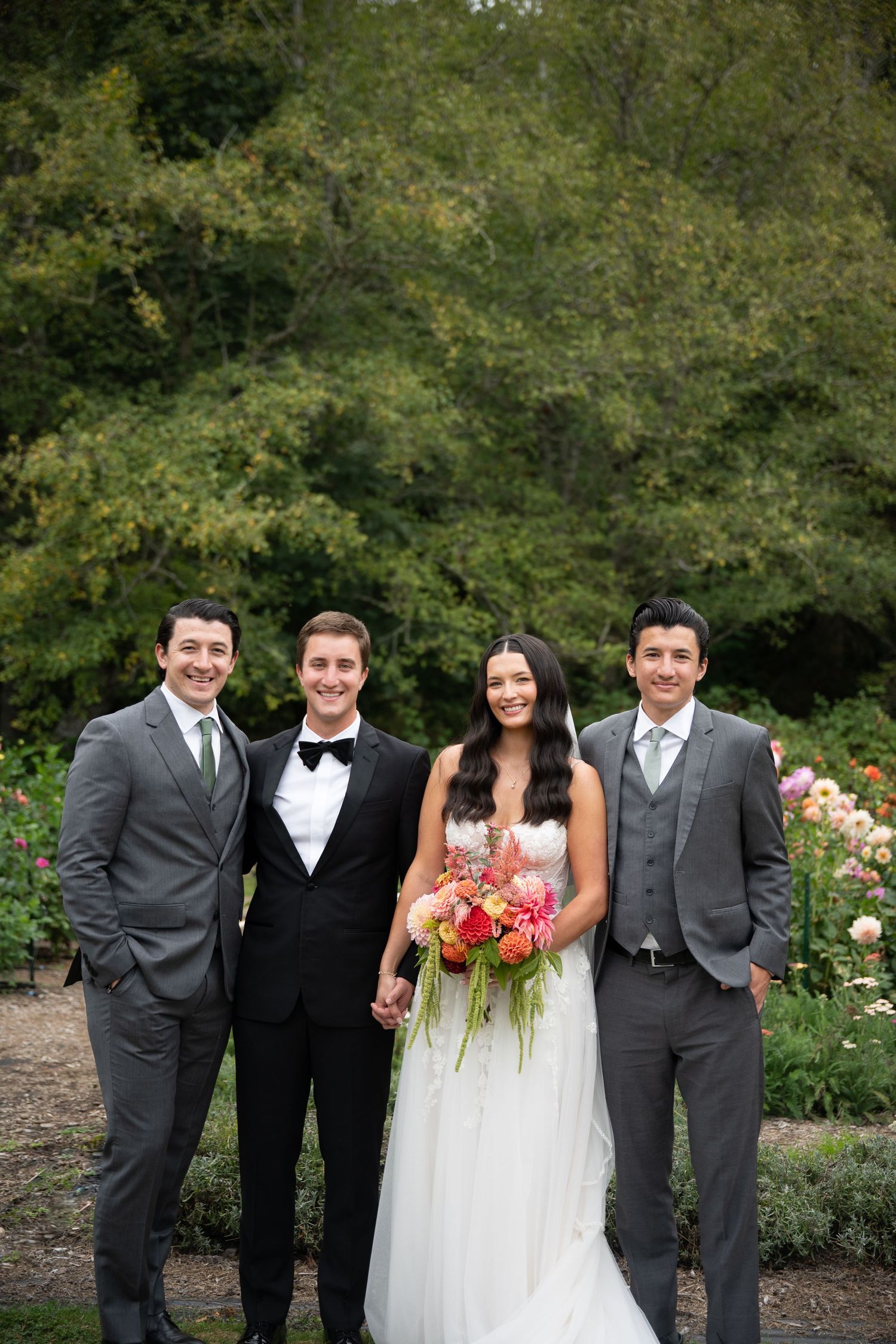 Bride holding colorful bouquet poses with groom and two groomsmen in gray suits outdoors.