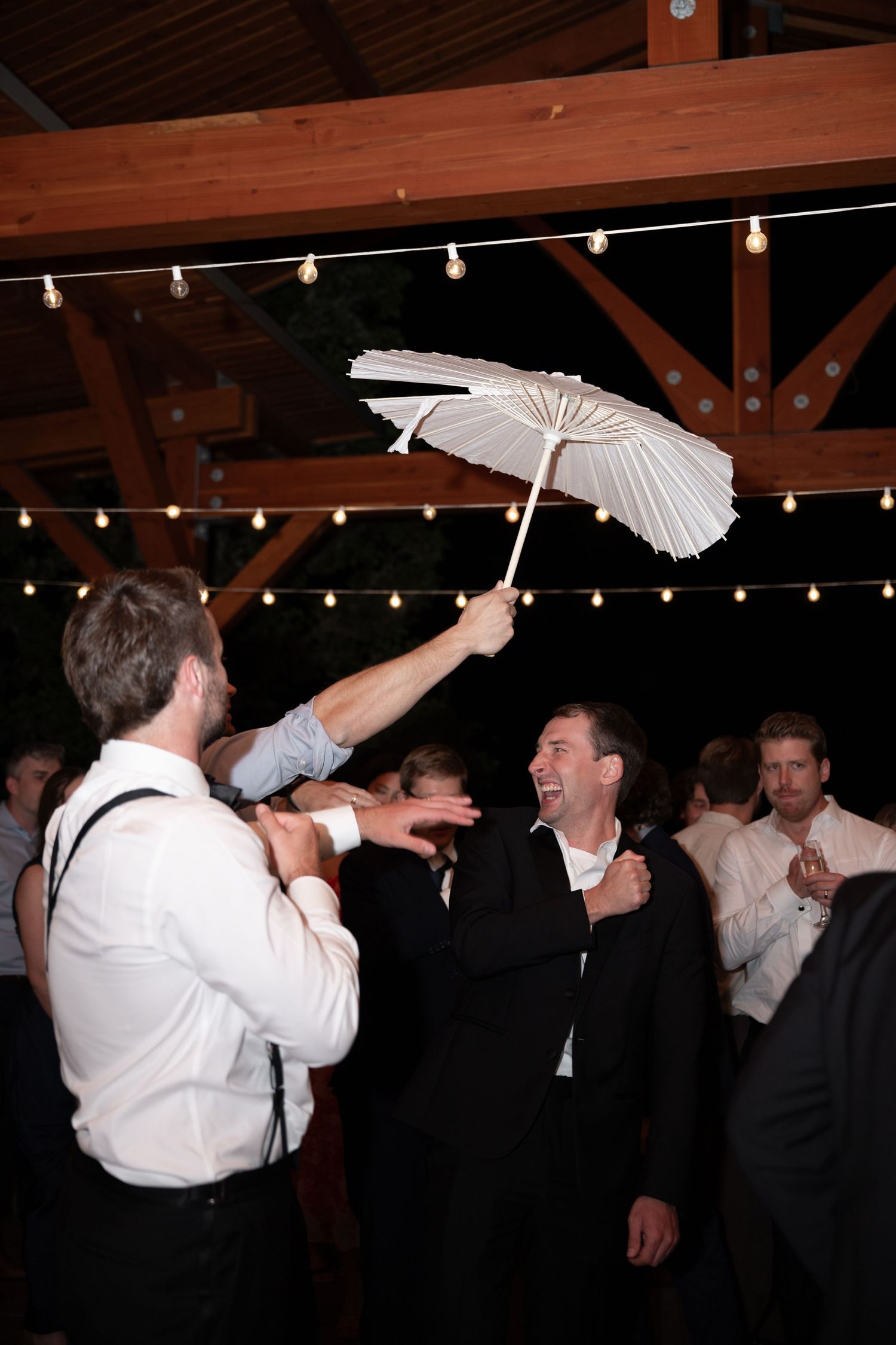 Guests laughing as man twirls white paper parasol at lively indoor wedding reception.