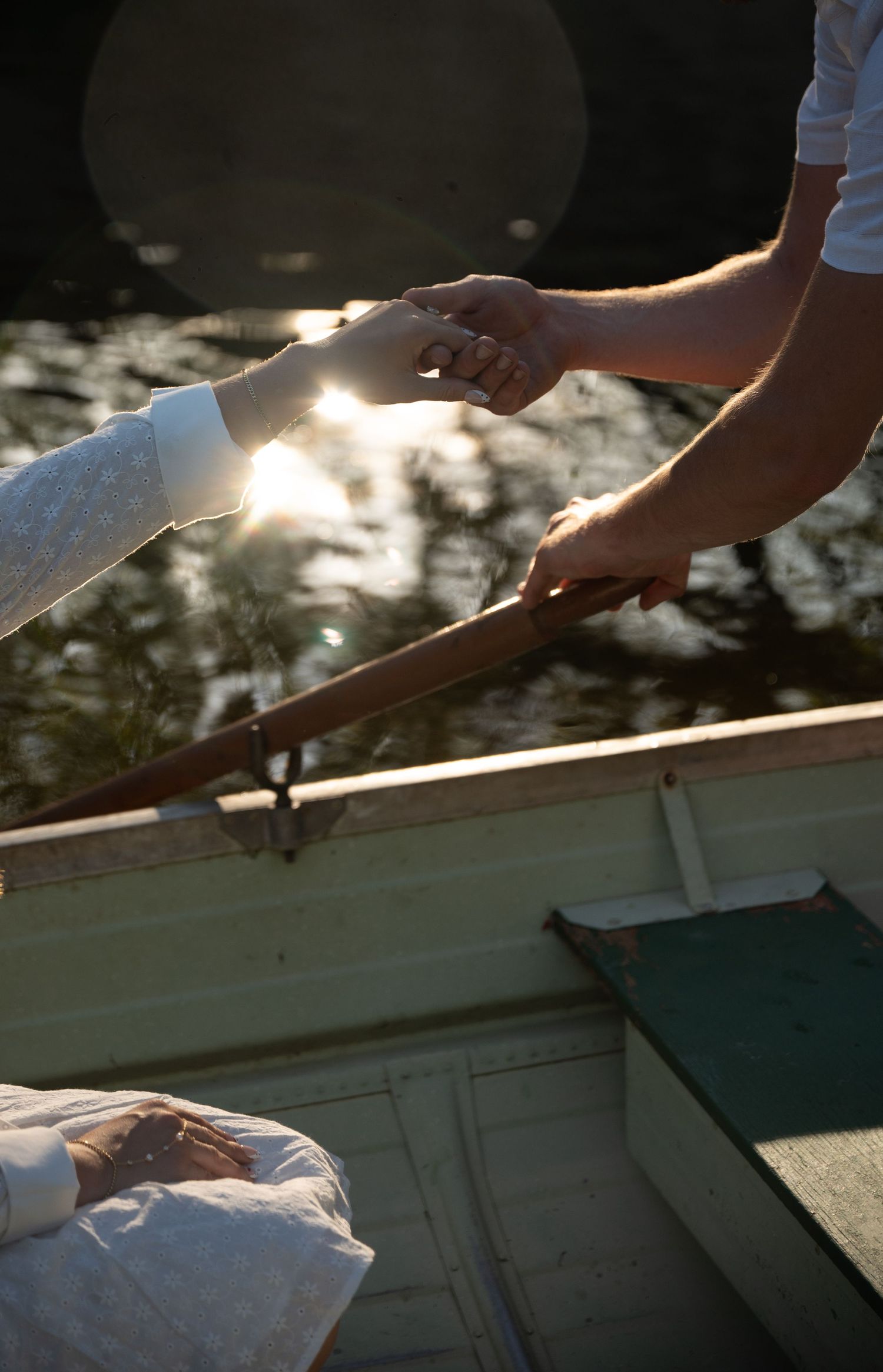 Two people holding hands over a wooden rowboat on a sunlit lake, with lens flare and water reflections.