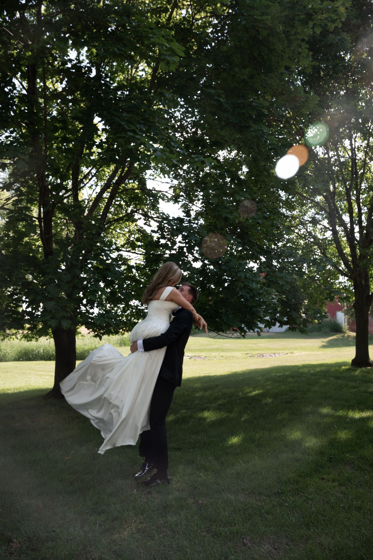 Groom lifting bride in white wedding gown in sunlit outdoor garden with tall green trees.