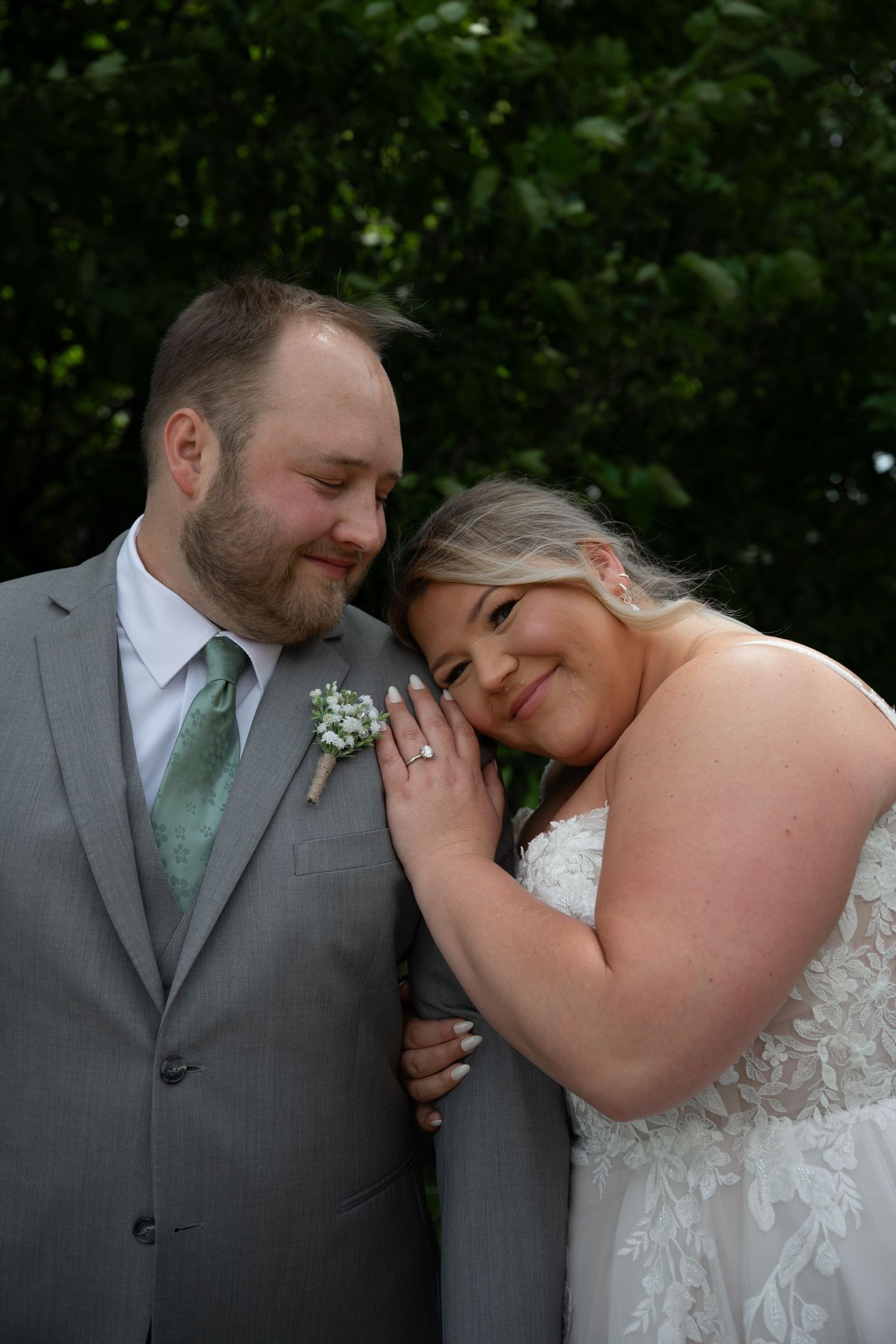 Bride leans head on groom's chest smiling, groom in gray suit with green tie, outdoor wedding portrait.