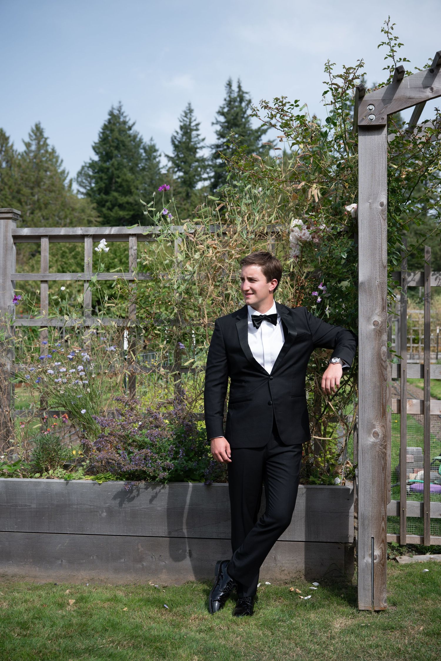 Groom in black tuxedo with bow tie leaning against wooden garden gate surrounded by lush greenery.