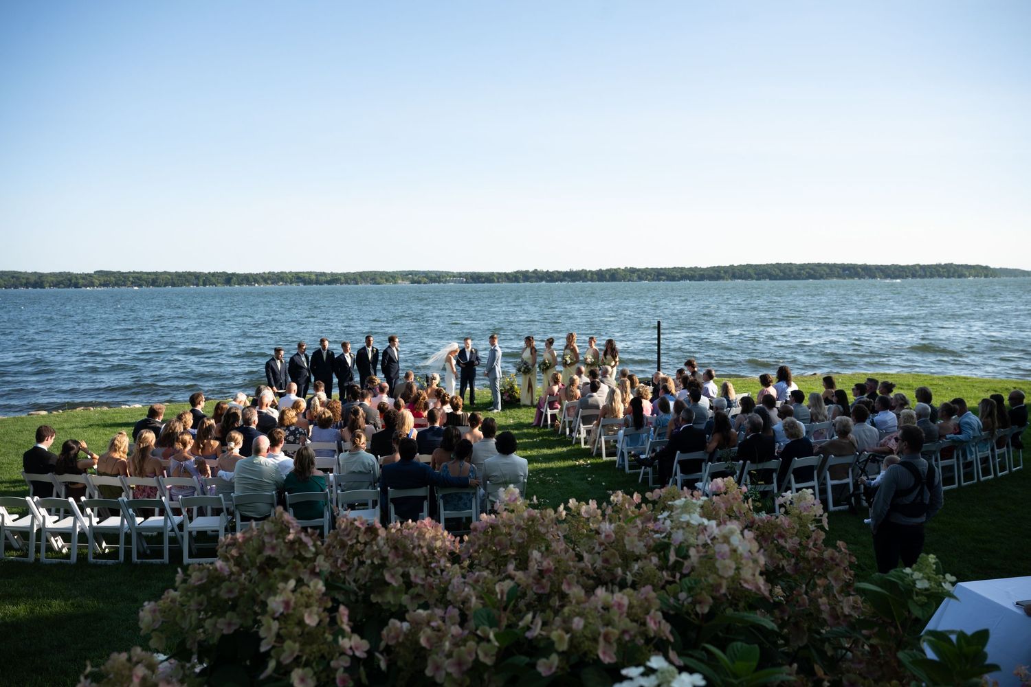 Outdoor waterfront wedding ceremony with guests seated on white chairs overlooking a serene lake under a clear blue sky.