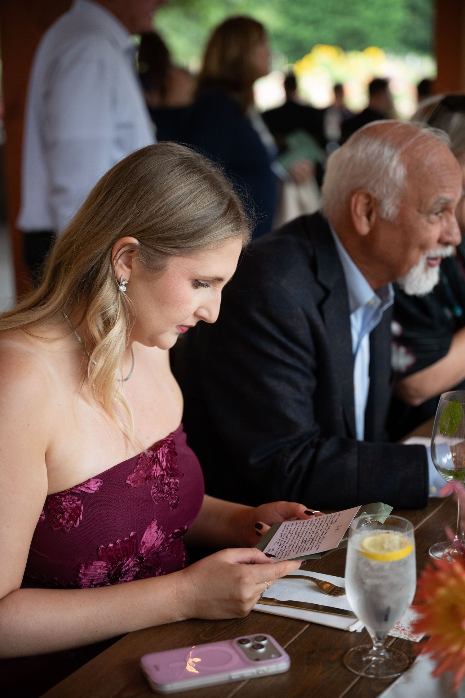Blonde wedding guest in burgundy floral dress reads menu card at outdoor reception table.