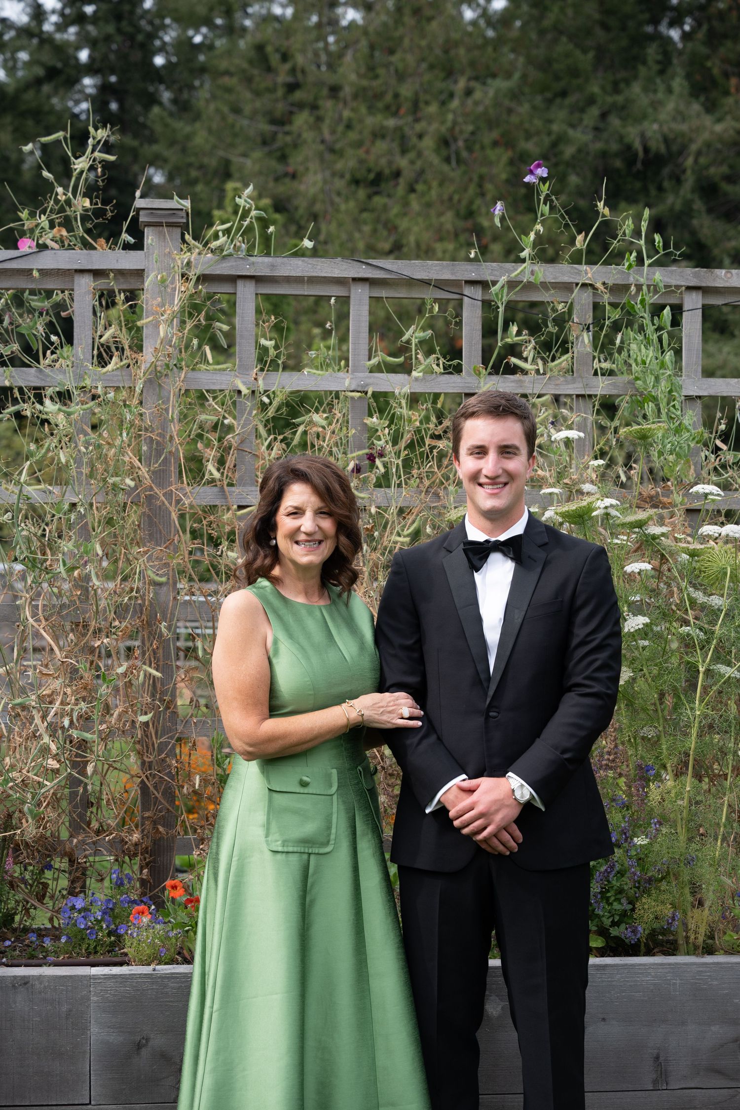 Woman in green gown and young man in tuxedo posing together in a garden setting.