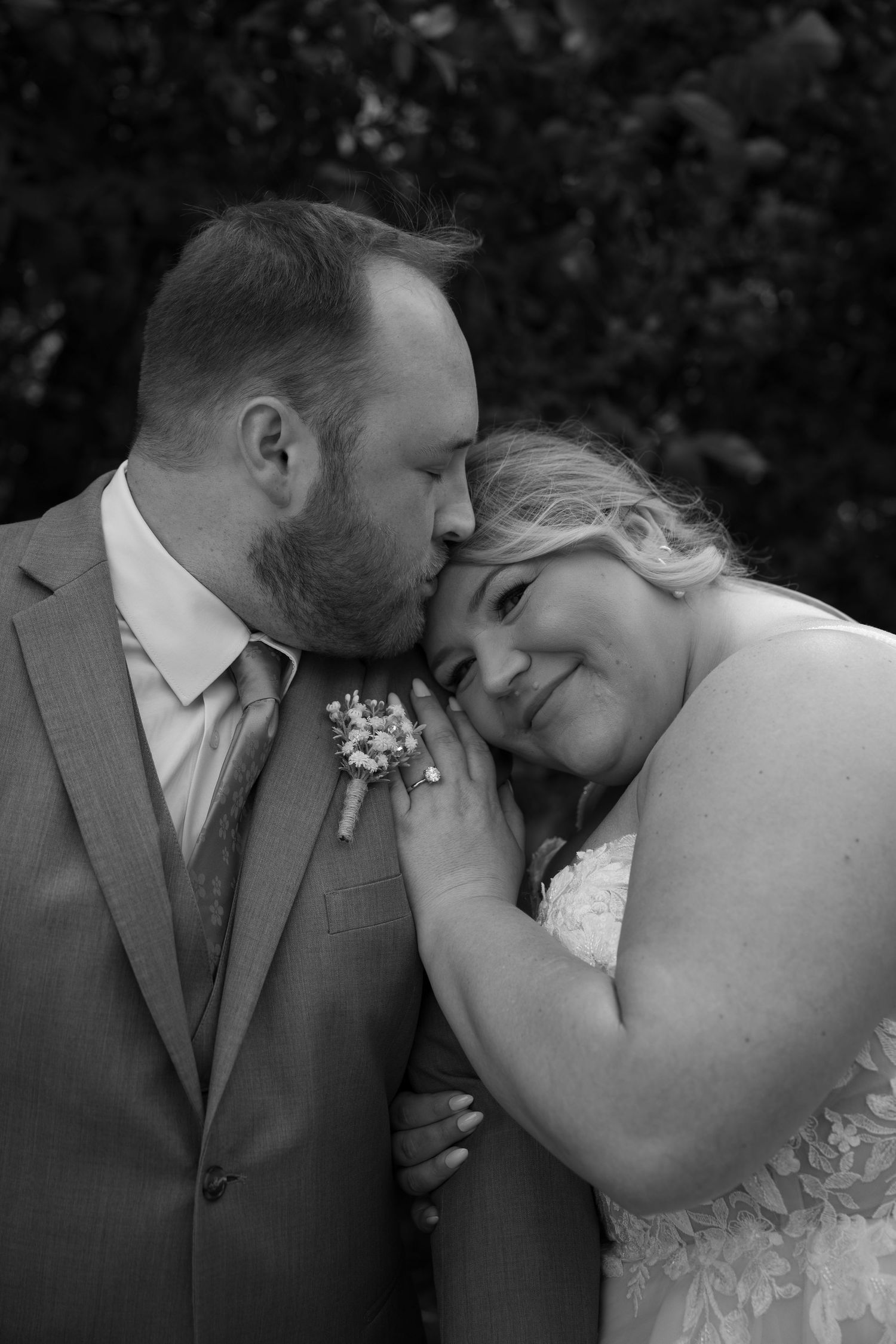 Black and white photo of groom kissing bride's forehead tenderly, both in wedding attire outdoors.