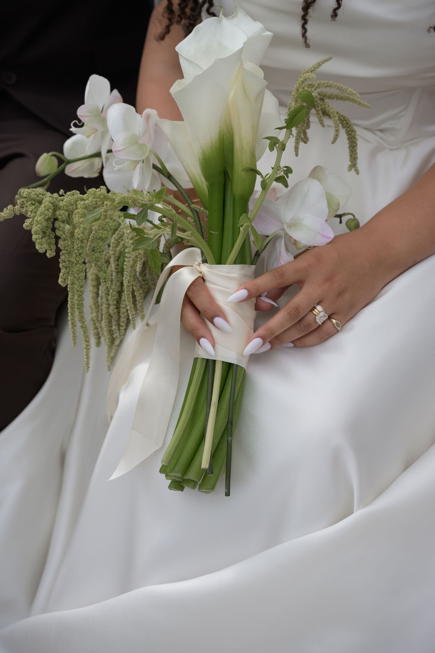 Bride in white dress holding elegant wedding bouquet of calla lilies and white flowers tied with satin ribbon, wearing diamond ring.