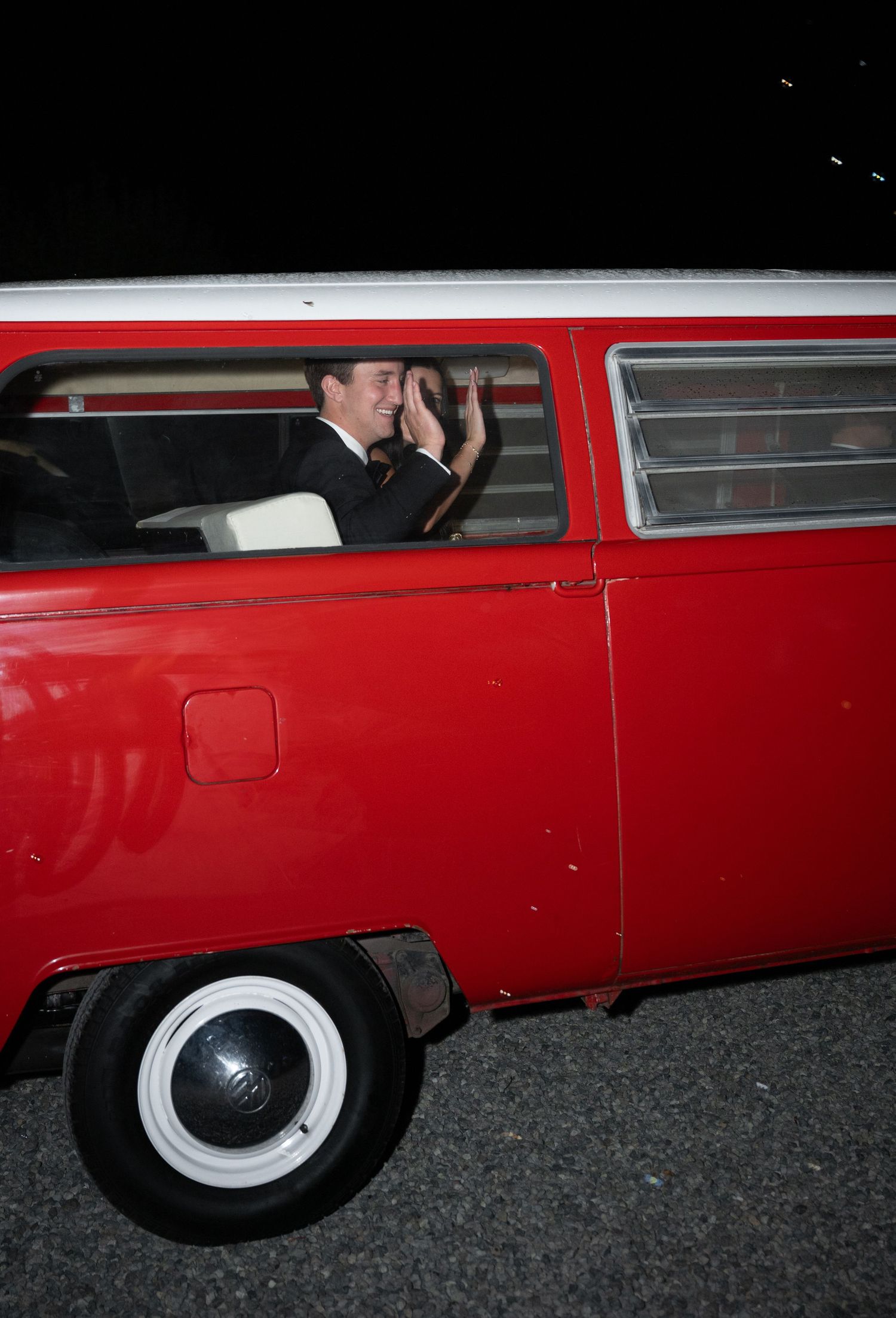 Groom in black tuxedo waves from a vintage red and white VW bus at night after wedding reception.