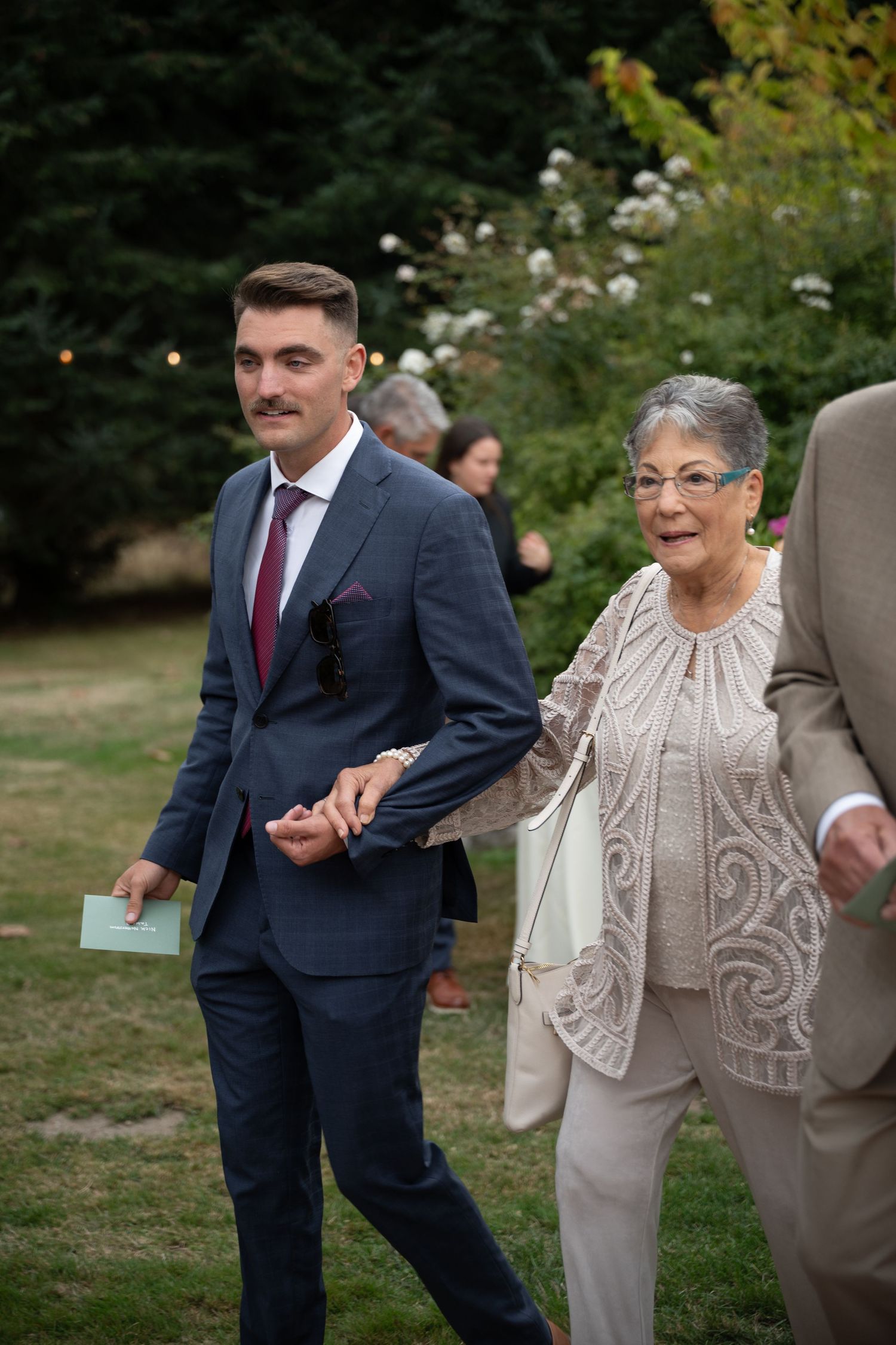 Young man in navy suit escorts elderly woman in beige lace dress at outdoor wedding ceremony.
