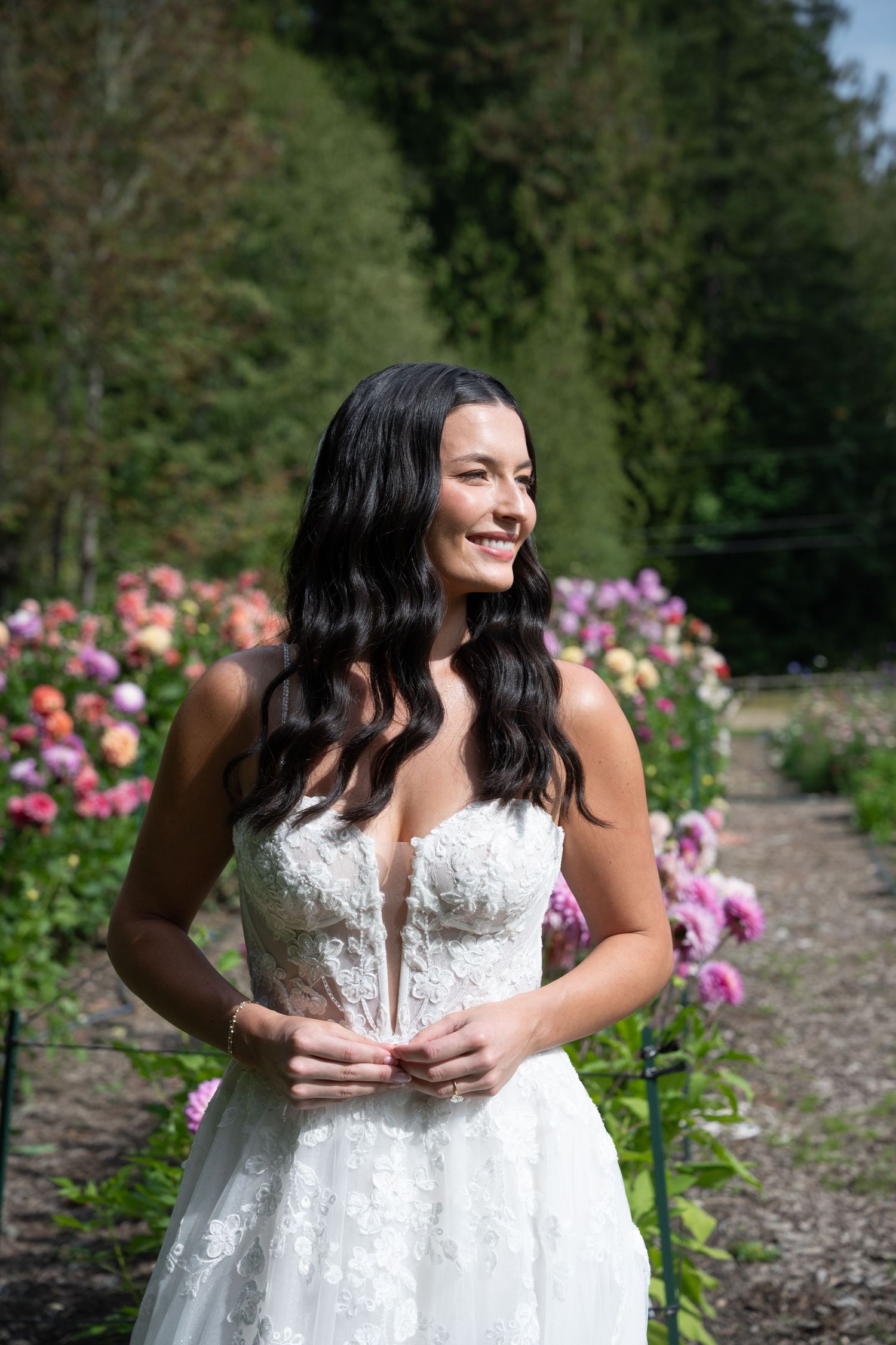 Smiling bride in a strapless lace wedding gown standing in a vibrant garden of pink and coral blooms on a sunny day.
