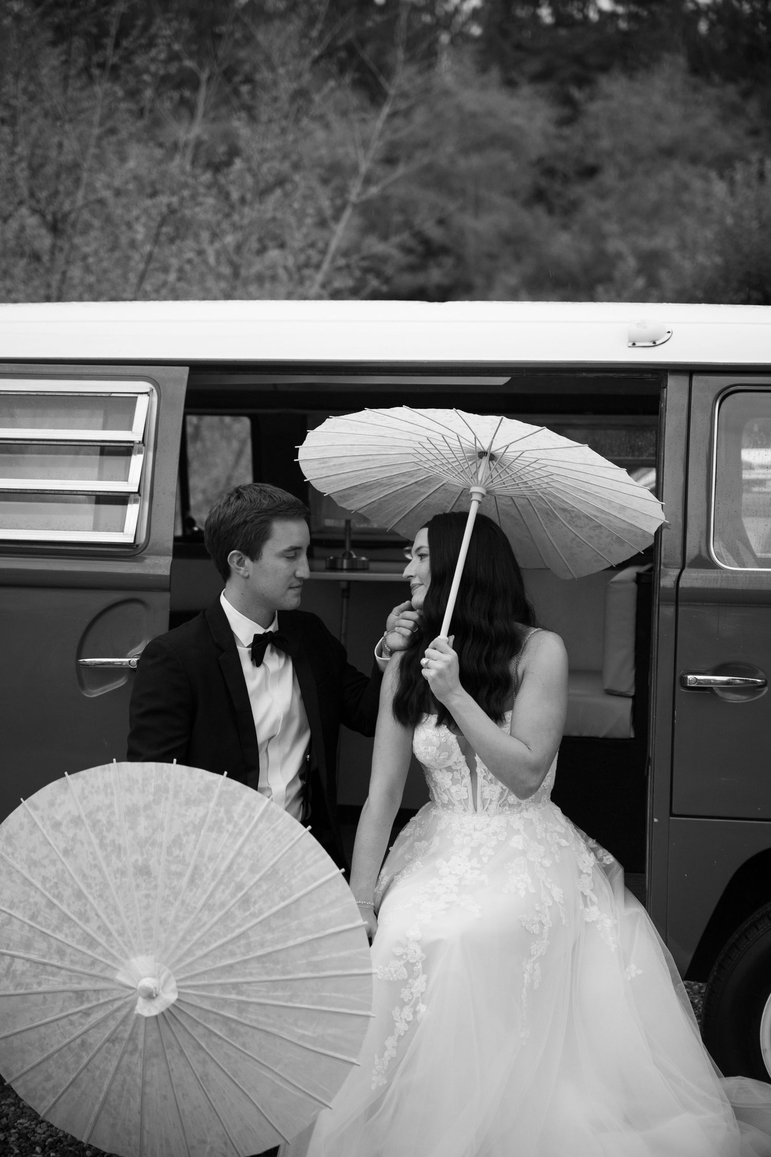 Black and white photo of a bride and groom holding parasols beside a vintage van on their wedding day.