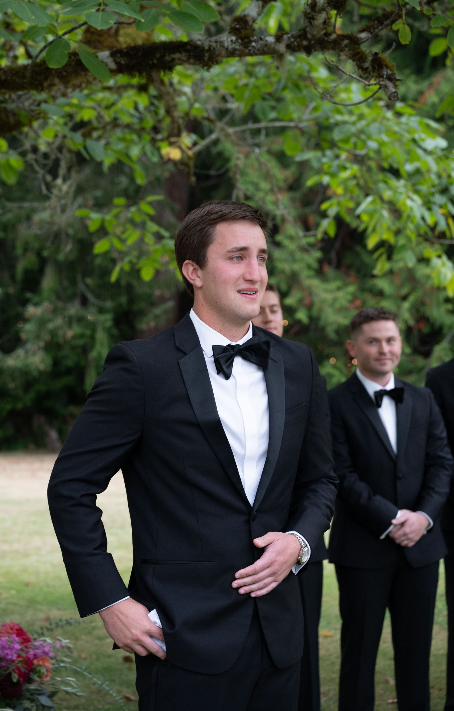 Groom in black tuxedo with bow tie smiling outdoors at wedding ceremony with groomsmen in background.