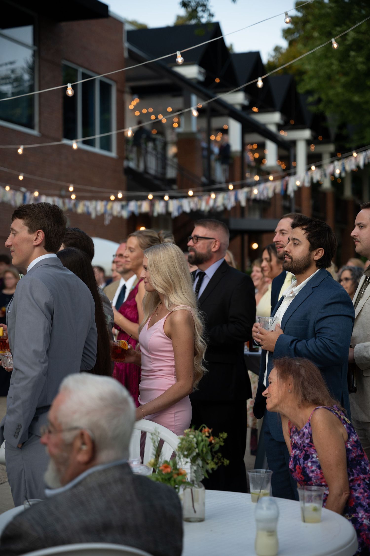 Guests mingle at an outdoor evening wedding reception with string lights and bunting decorations in an urban setting.